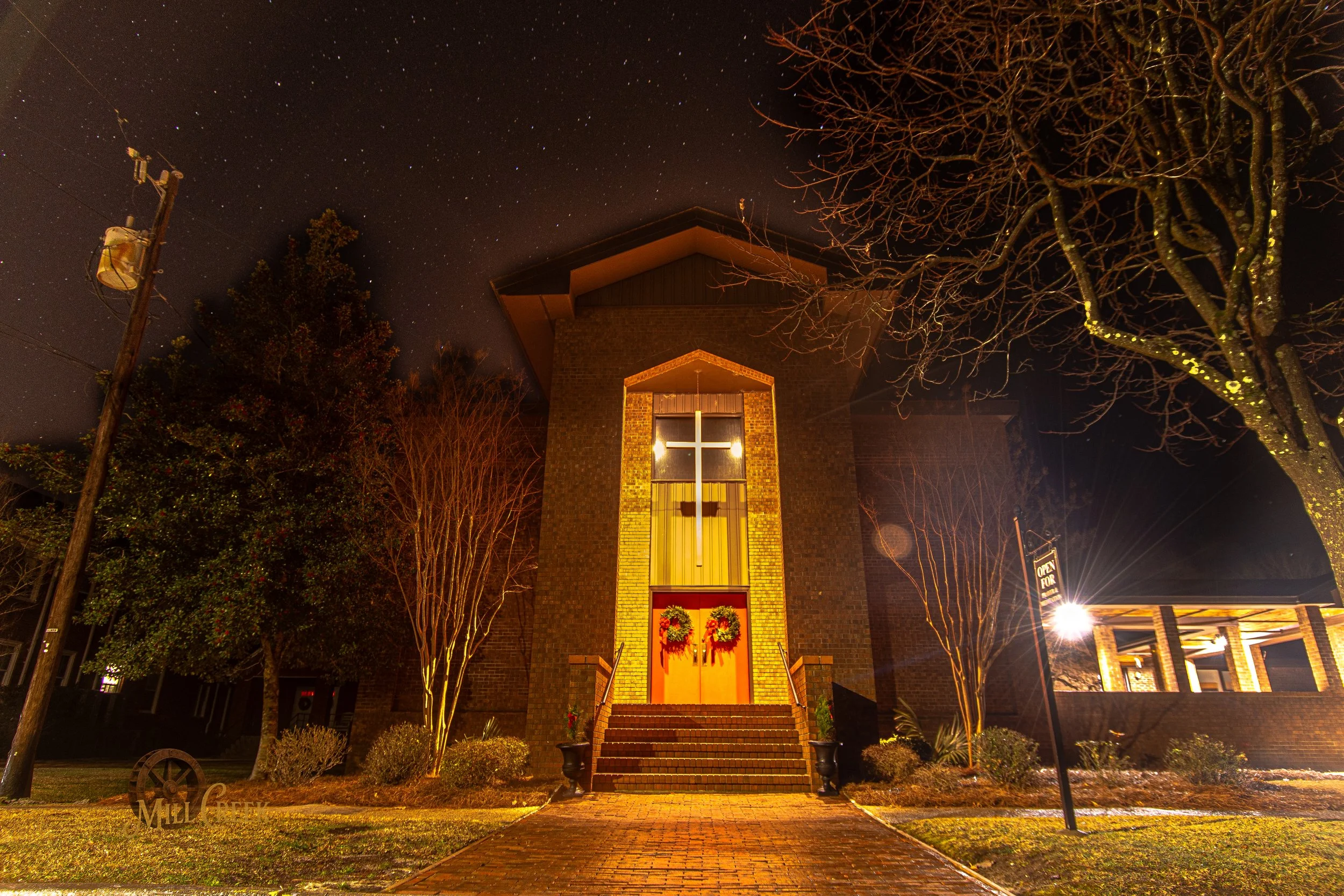 Nighttime view of a brick church with a large illuminated cross above the doorway, decorated with Christmas wreaths, and surrounded by trees and a lit pathway.