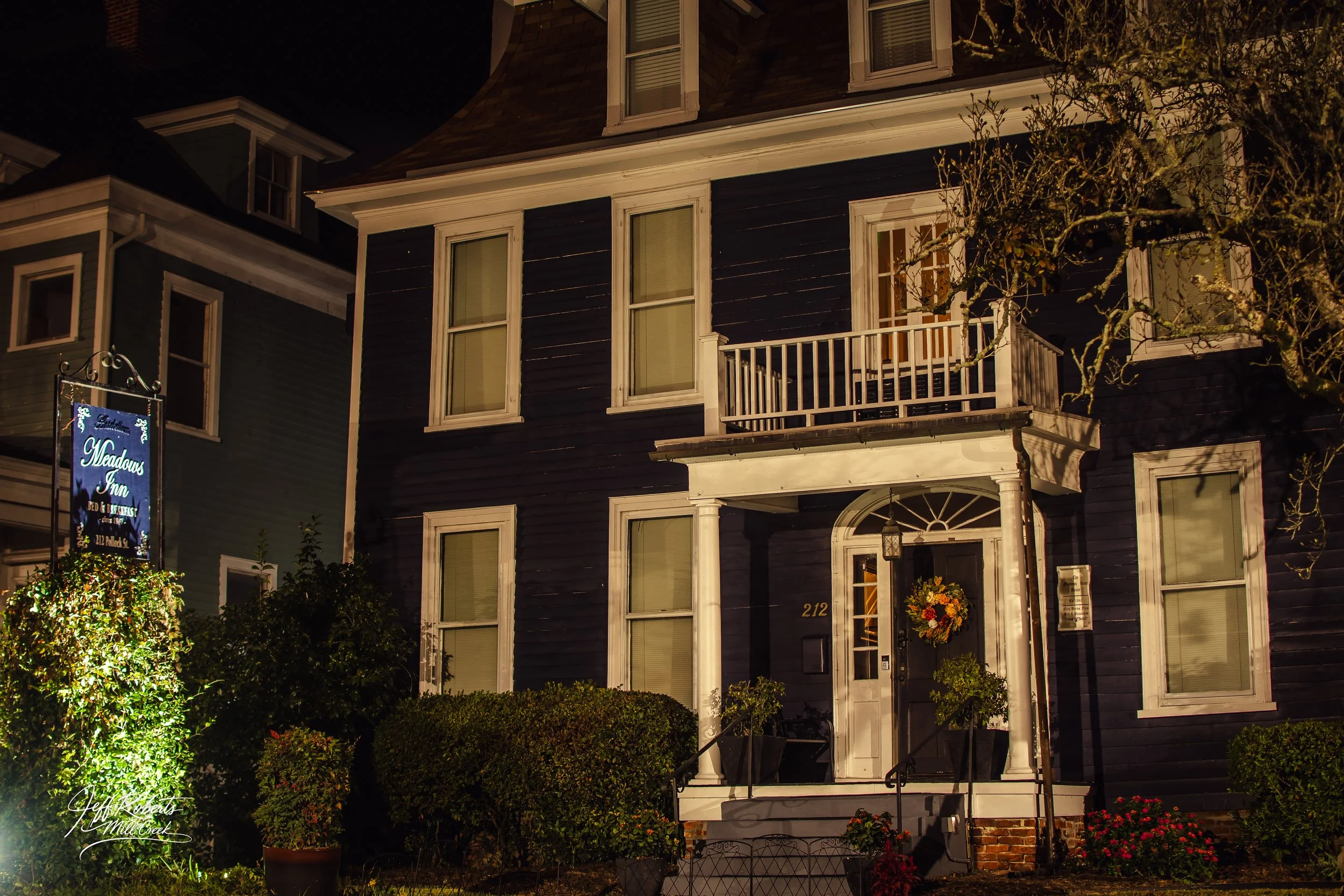 A dark blue three-story house with white trim and a small balcony on the second floor, decorated with a wreath on the front door, with a sign reading 'Meadows Inn' on the left side, surrounded by bushes and plants at night.