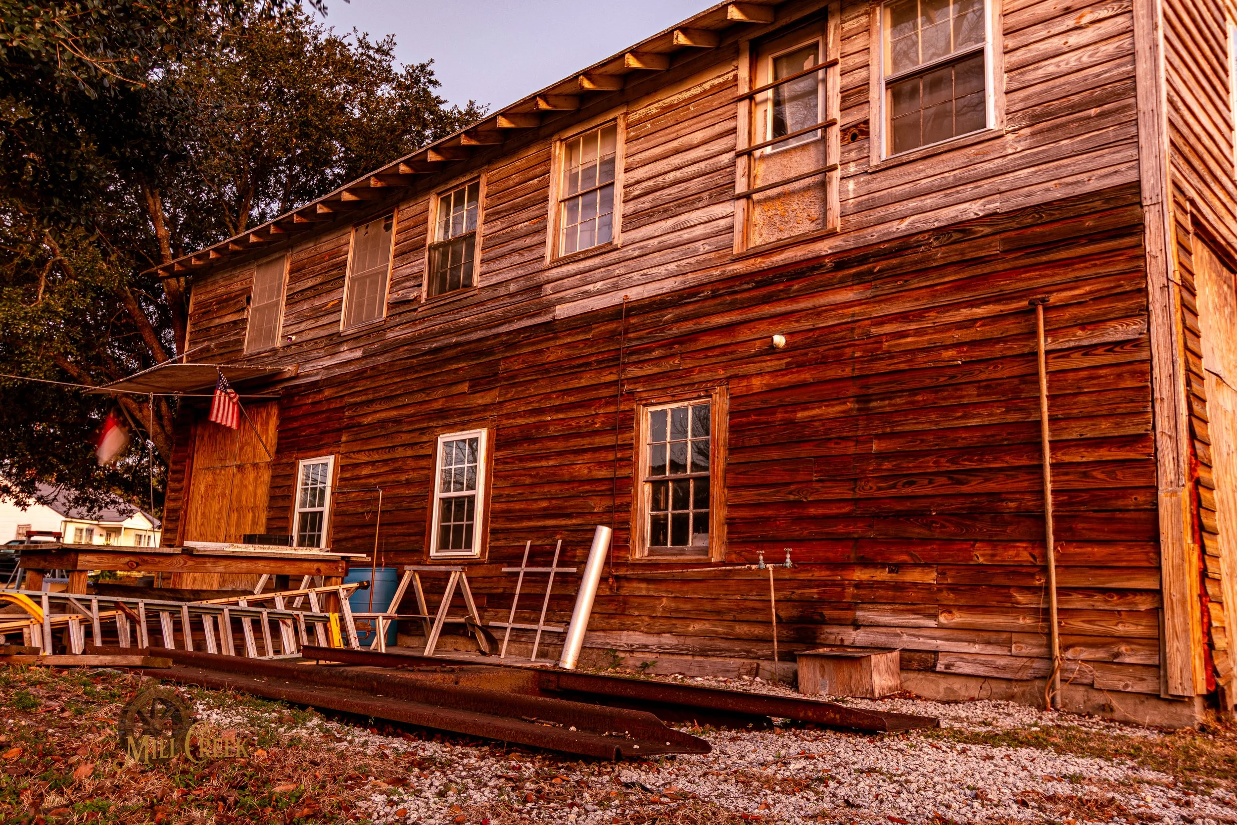 A rustic wooden building with weathered horizontal planks, multiple windows, and a small porch. There are American flags and various construction or maintenance items outside.