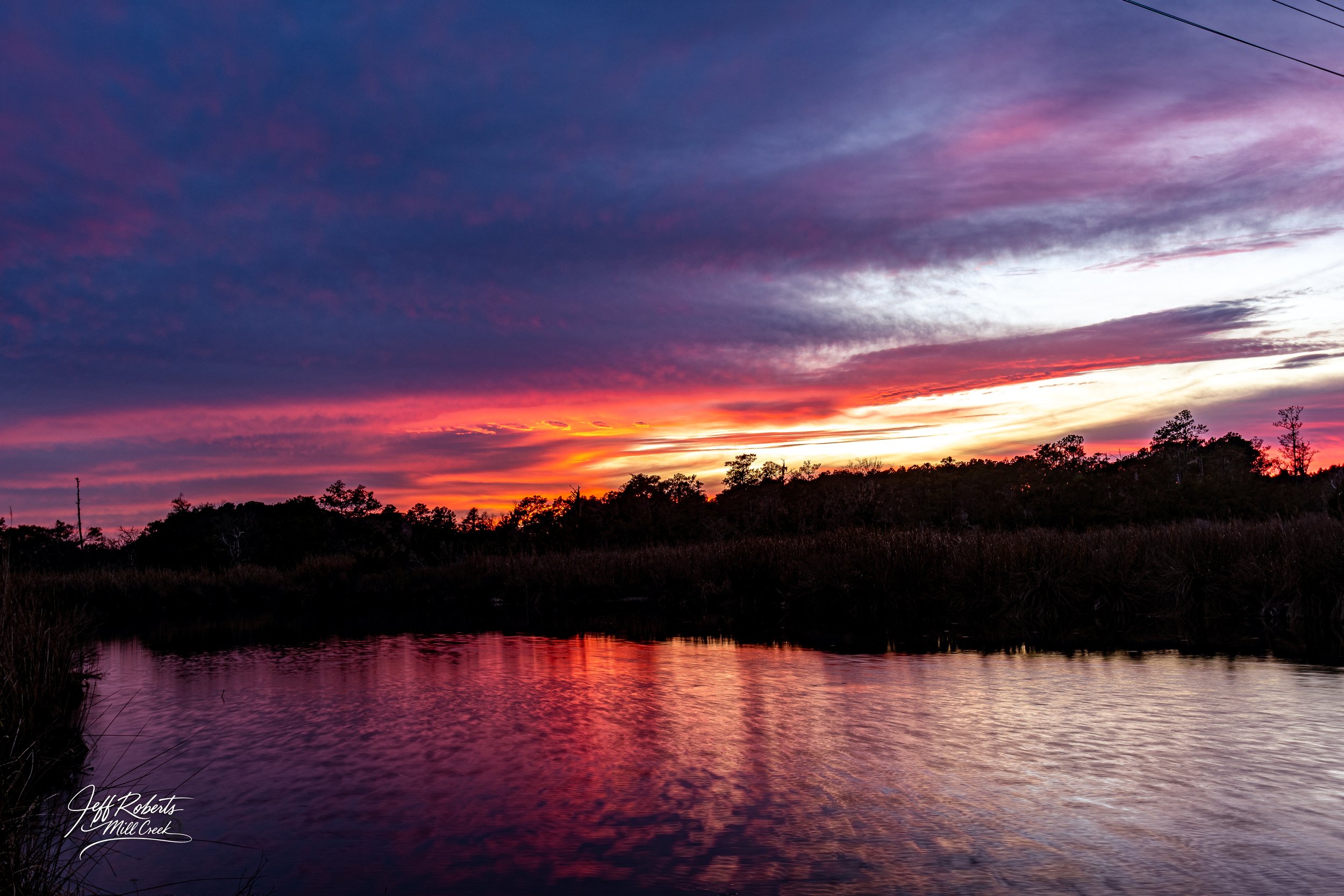 Sunset over a river with trees and bushes along the shoreline, colorful clouds in the sky reflecting in the water.