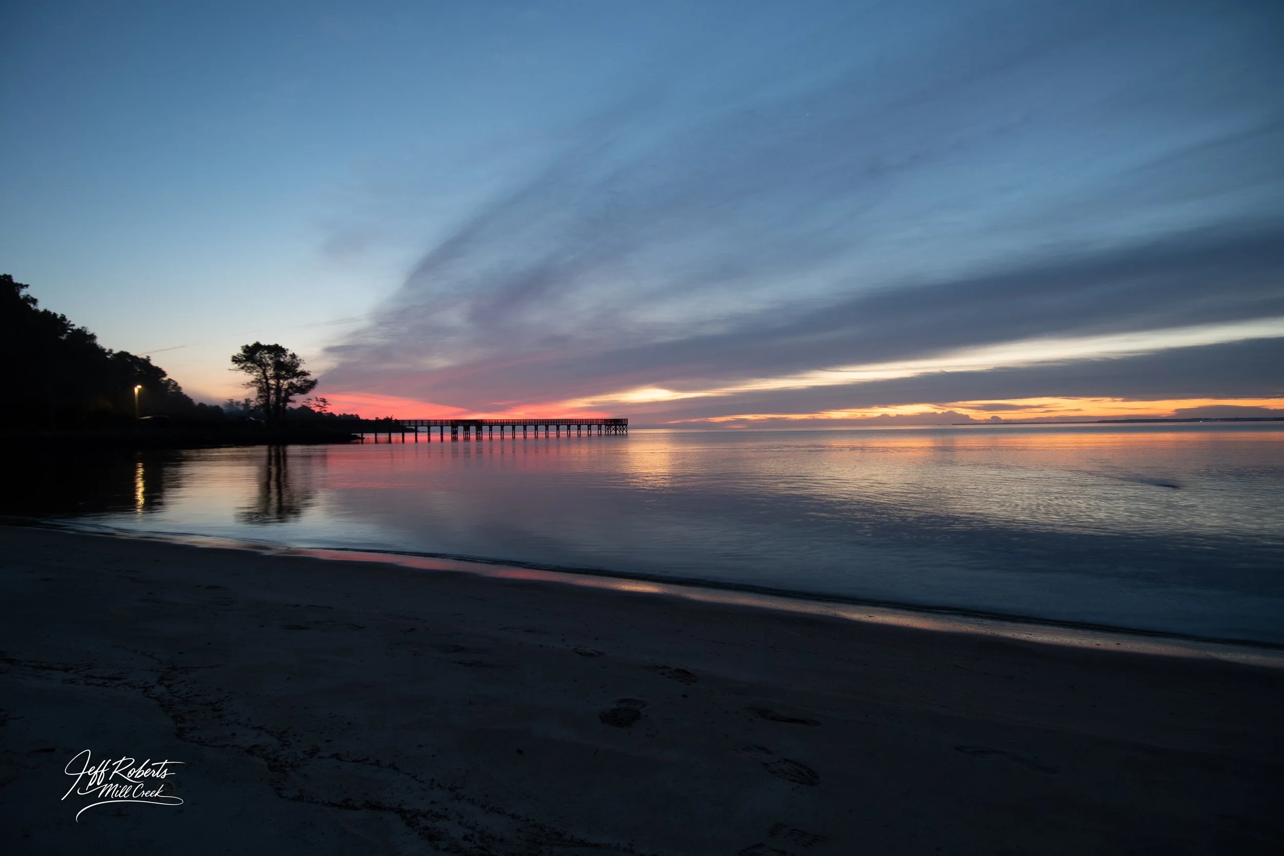Sunset over a calm body of water with a pier extending into the distance, silhouette of trees on the left, and a partly cloudy sky with streaks of orange, pink, and blue.