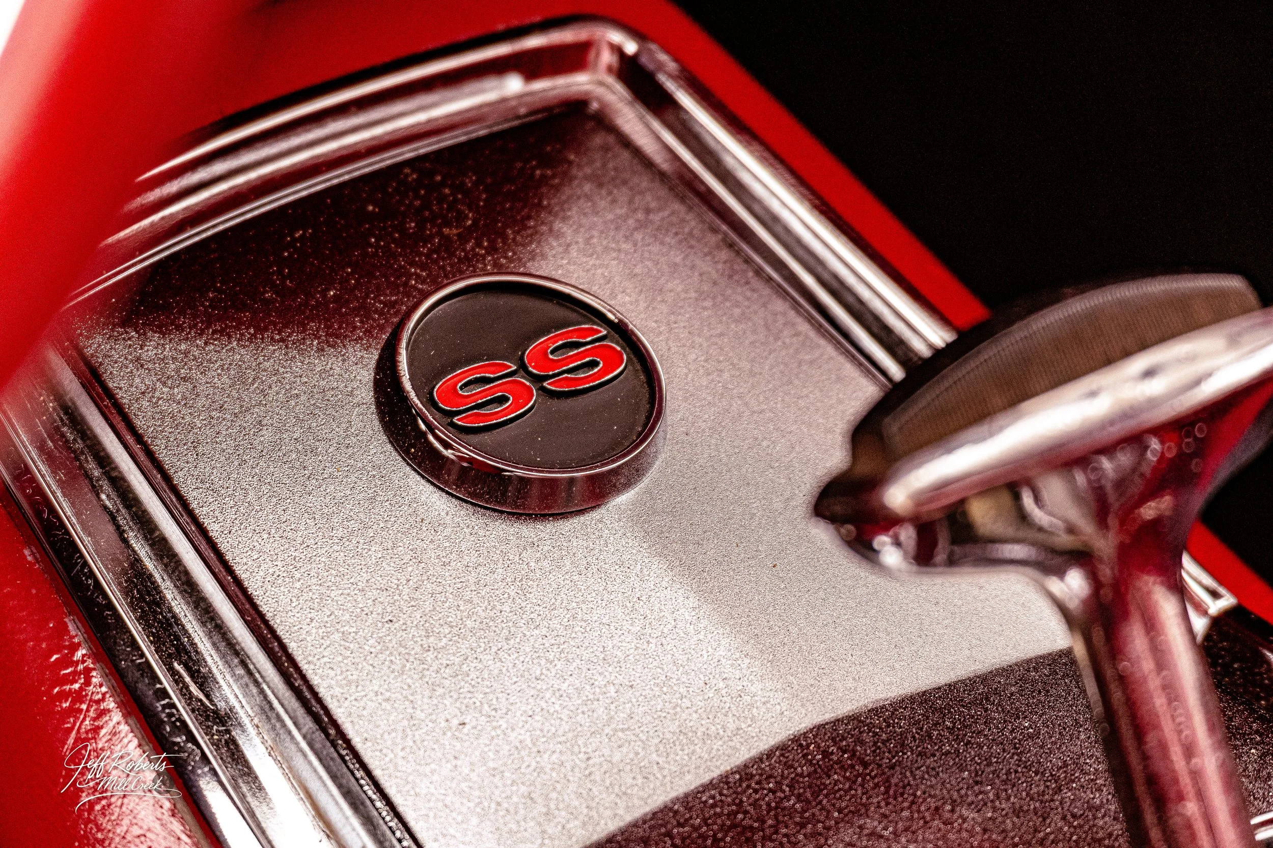 Close-up of a vintage car dashboard featuring a black circular emblem with red letters 'SS' and a metallic textured surface, with part of a steering wheel on the right.