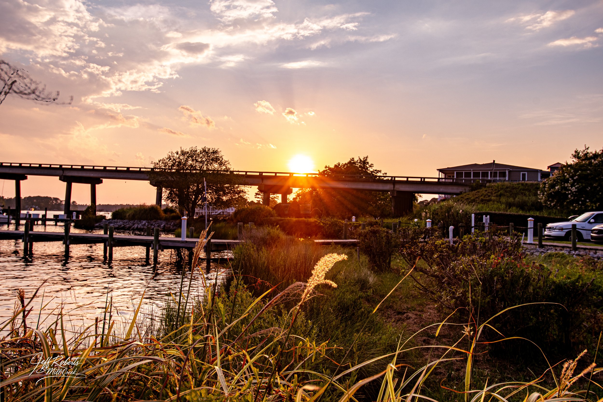 Sunset over a waterfront with a bridge, houses, trees, grass, and parked cars in the background.