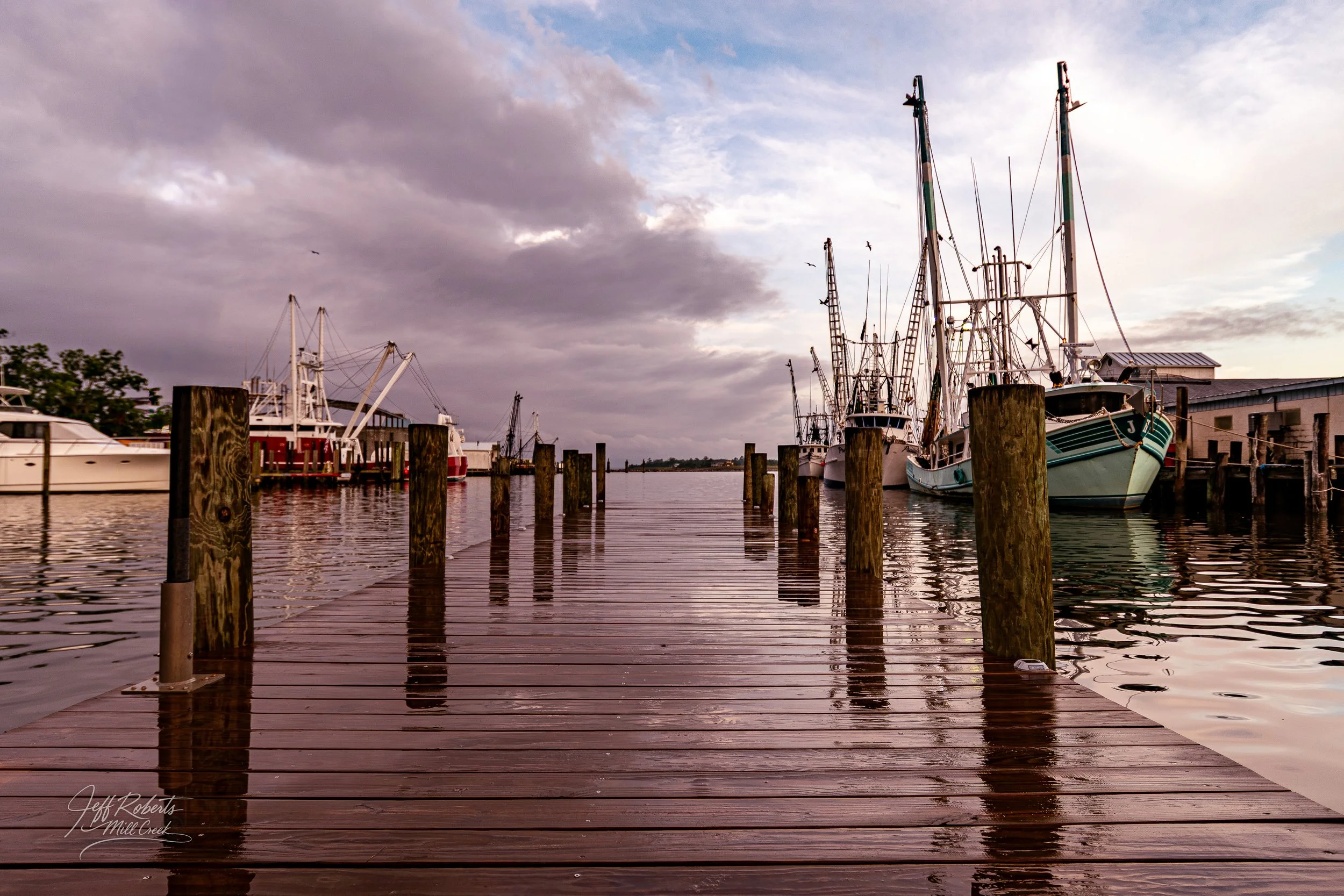 A wooden dock extending into a harbor with boats and yachts docked on either side, under a cloudy sky at dusk.