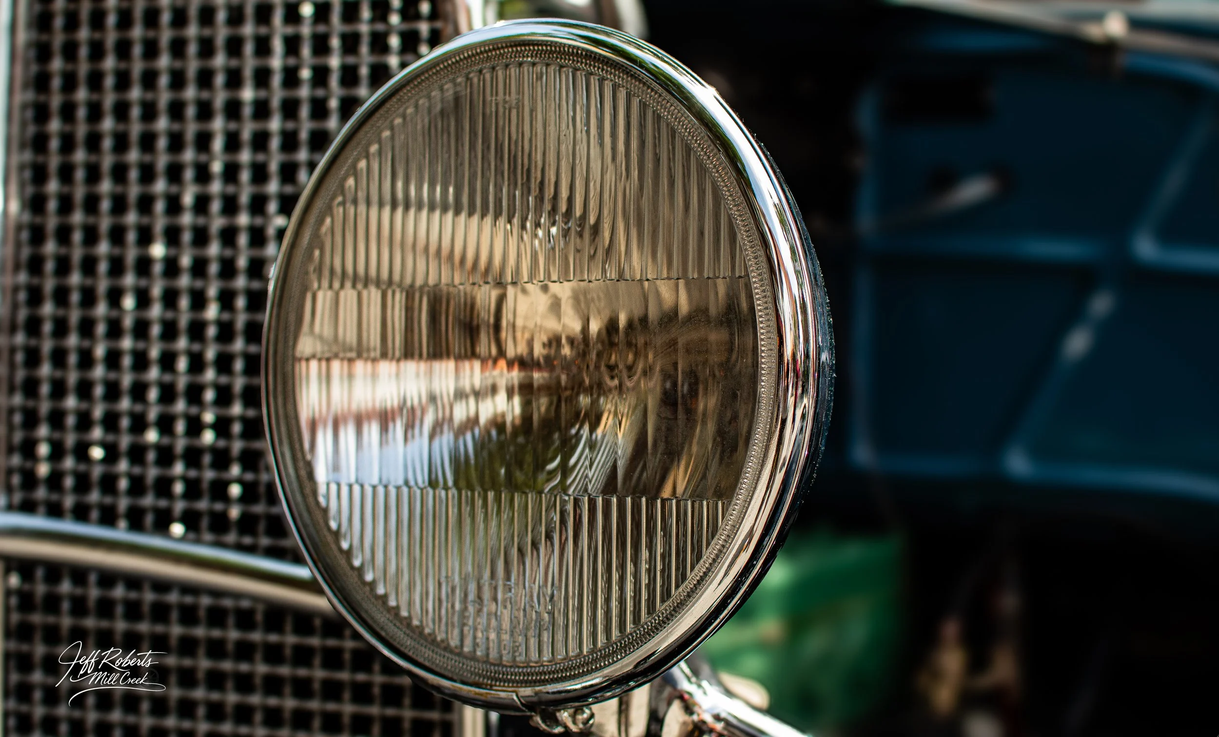 Close-up of a vintage car headlight with chrome rim and a textured glass lens, with the front grille and body of the car partially visible in the background.