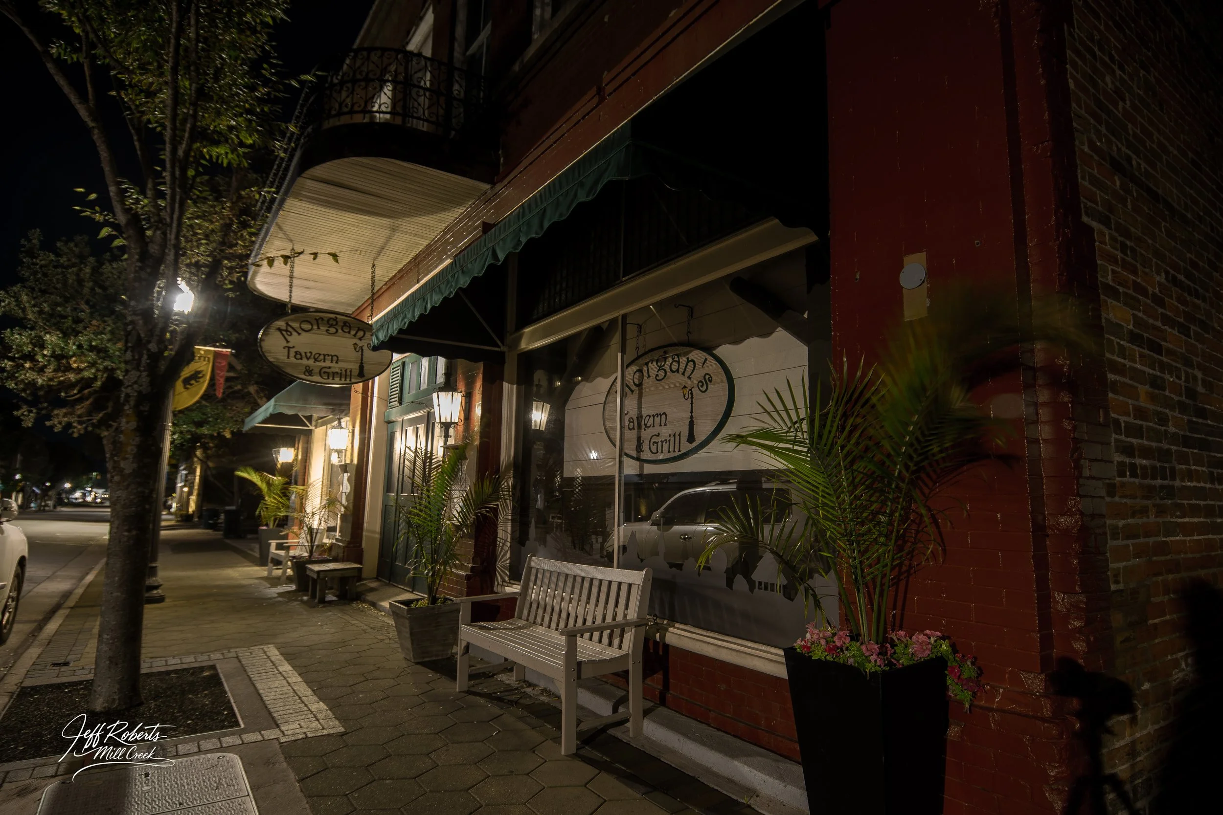 Nighttime street view of Morgan's Tavern & Grill storefront with illuminated sign, white bench, potted plants, and brick building exterior.