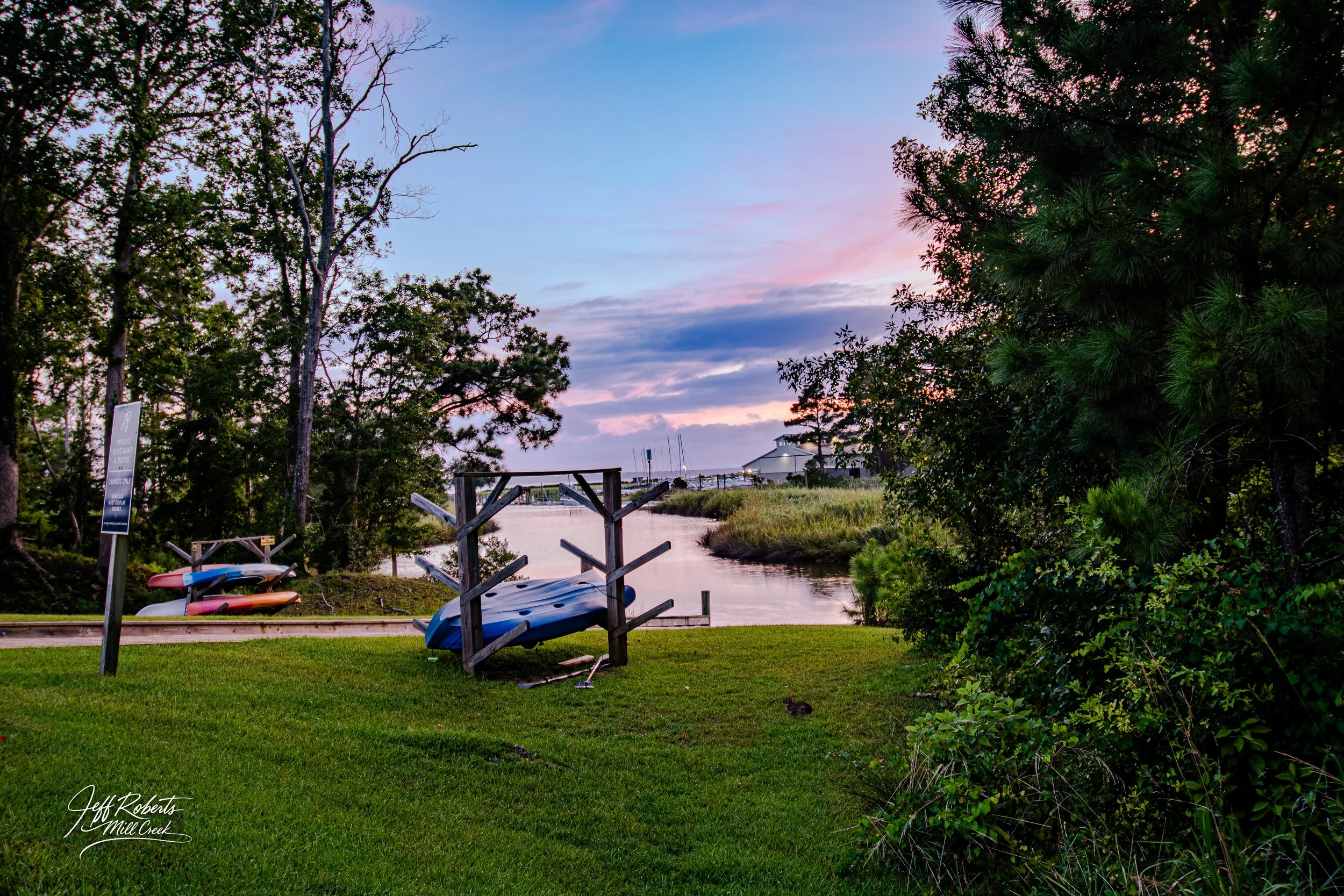A peaceful riverside scene at sunset with a grassy area, kayaks on a rack, trees, and a building in the distance under a colorful sky.