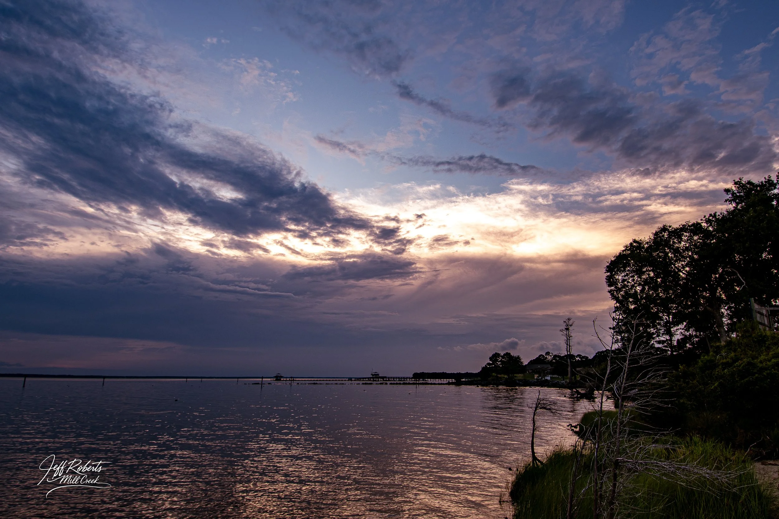 A scenic view of a body of water during sunset with a colorful sky filled with clouds, trees on the shoreline, and a calm reflective surface.