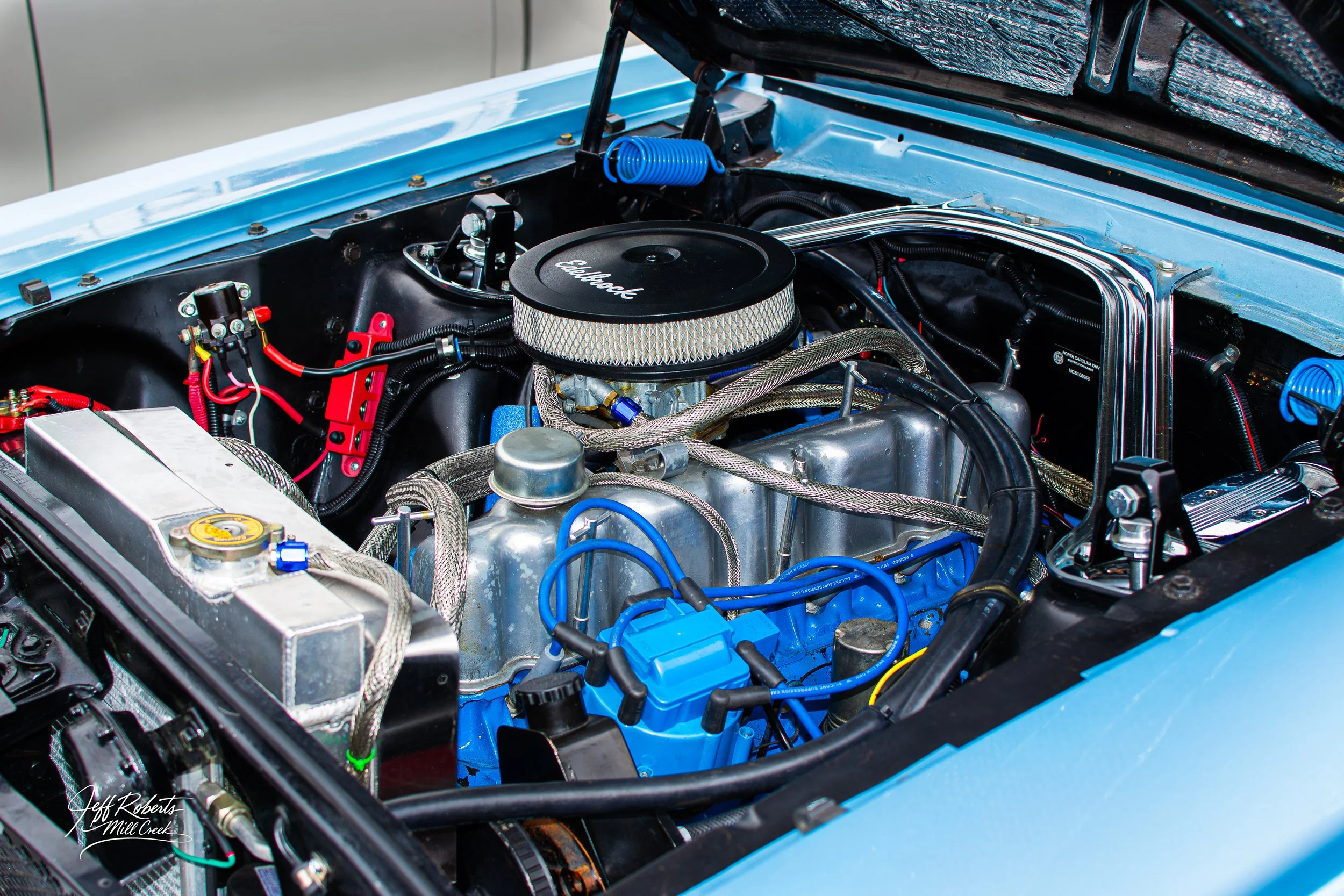 Close-up of a classic car engine with various metallic and colored components, including a black Edelbrock air filter, bright blue spark plug wires, and a silver metallic radiator.