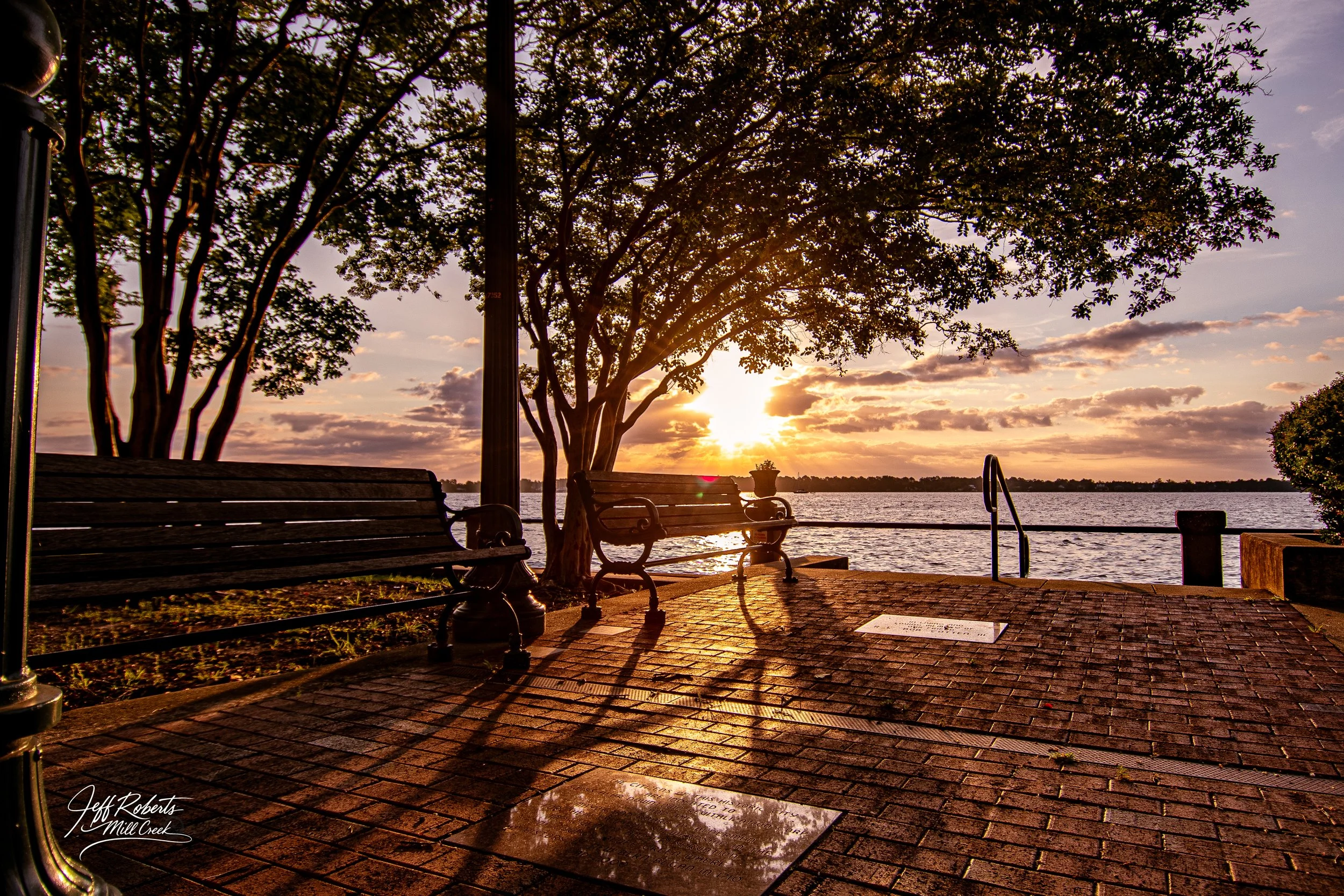 A lakeside scene at sunset with two benches on a brick patio, silhouetted trees, and water reflecting the sunset sky.