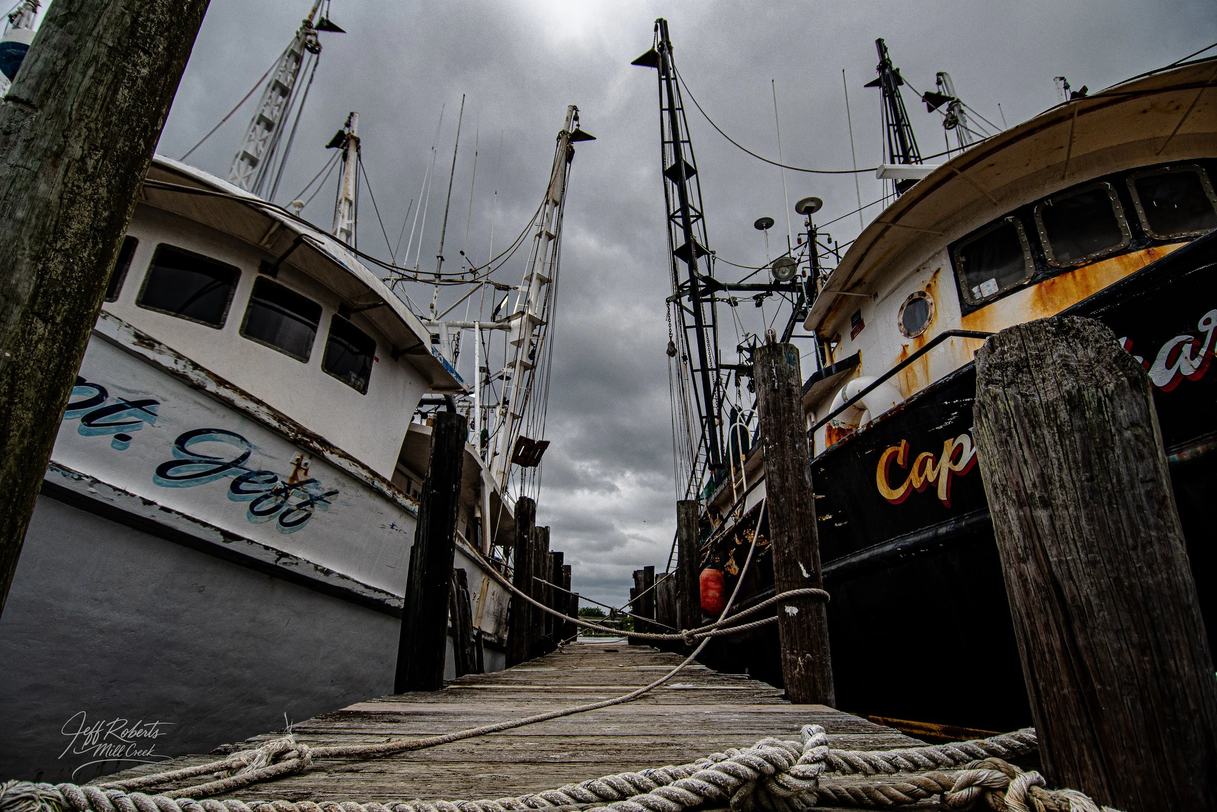 Two boats docked at a wooden pier under a cloudy sky, with ropes and wooden posts in the foreground.