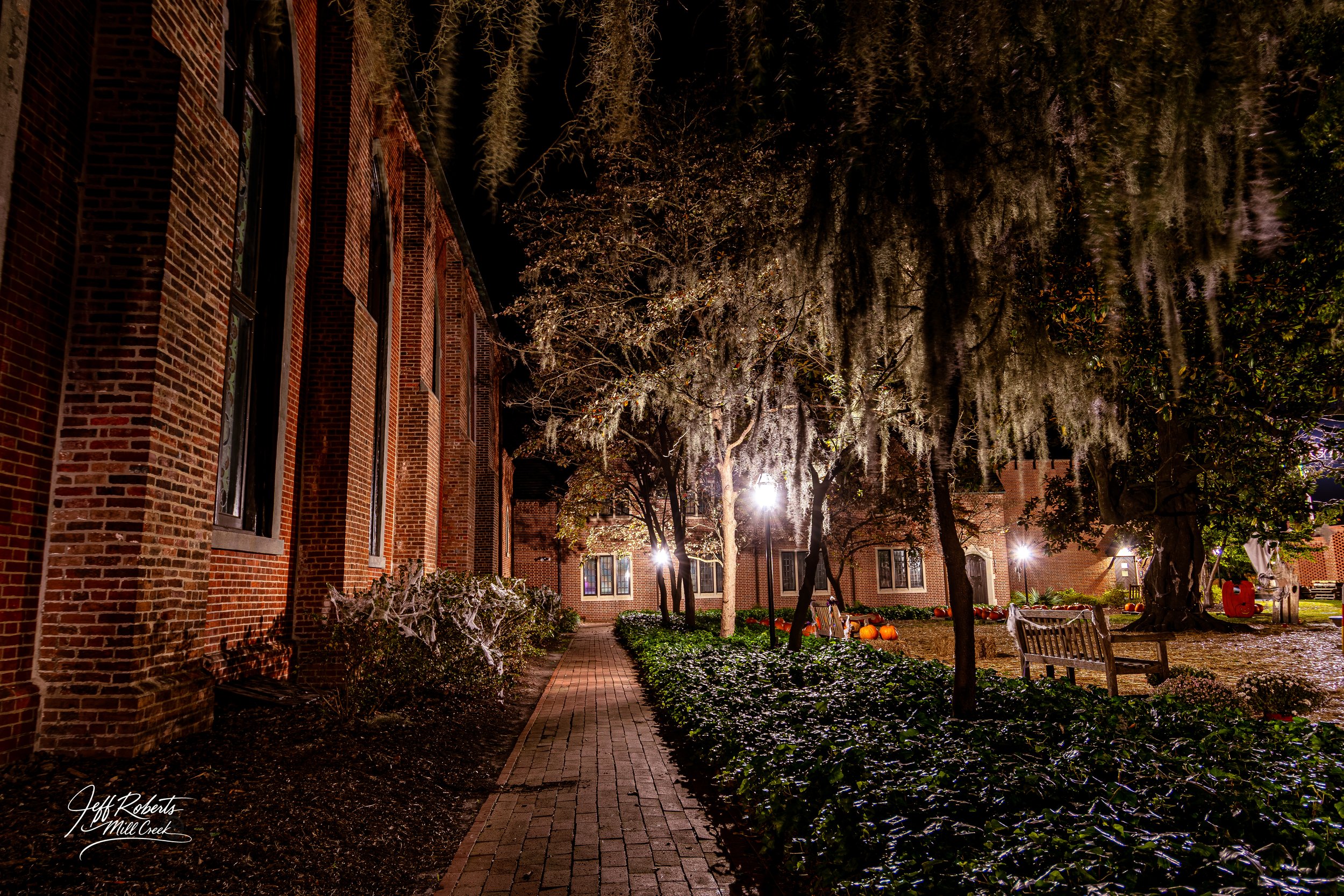 Night view of brick building with windows, lit pathway, trees, benches, and pumpkins.