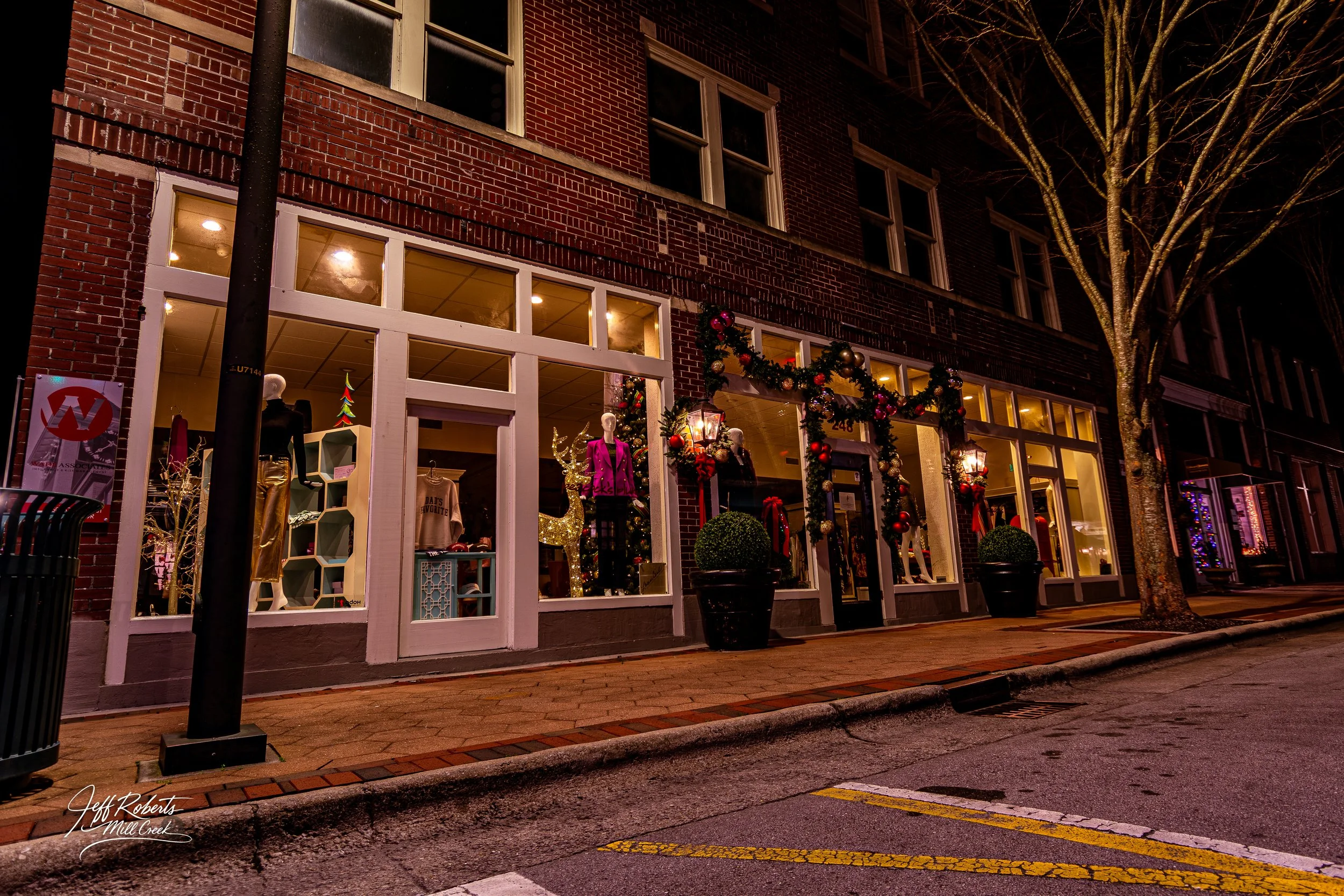 Store window decorated with Christmas garland, ornaments, and mannequins in holiday clothing, across a brick sidewalk at night.