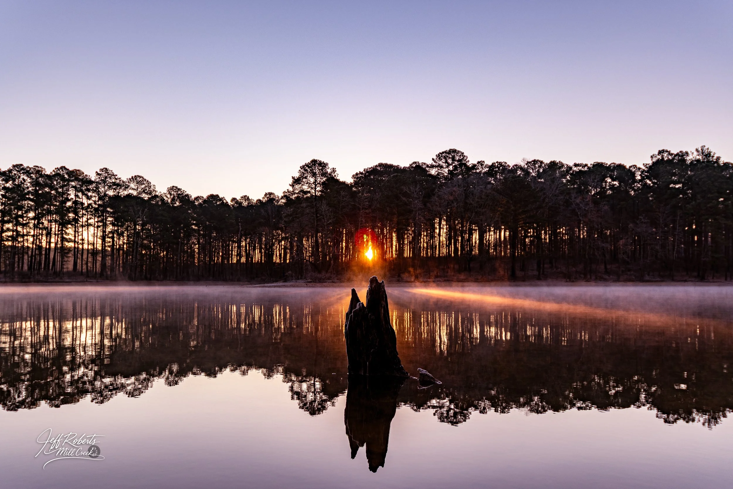 A serene lake at sunrise with fog near the water surface, trees lining the horizon, and the sun just peeking through the trees, casting a warm glow.