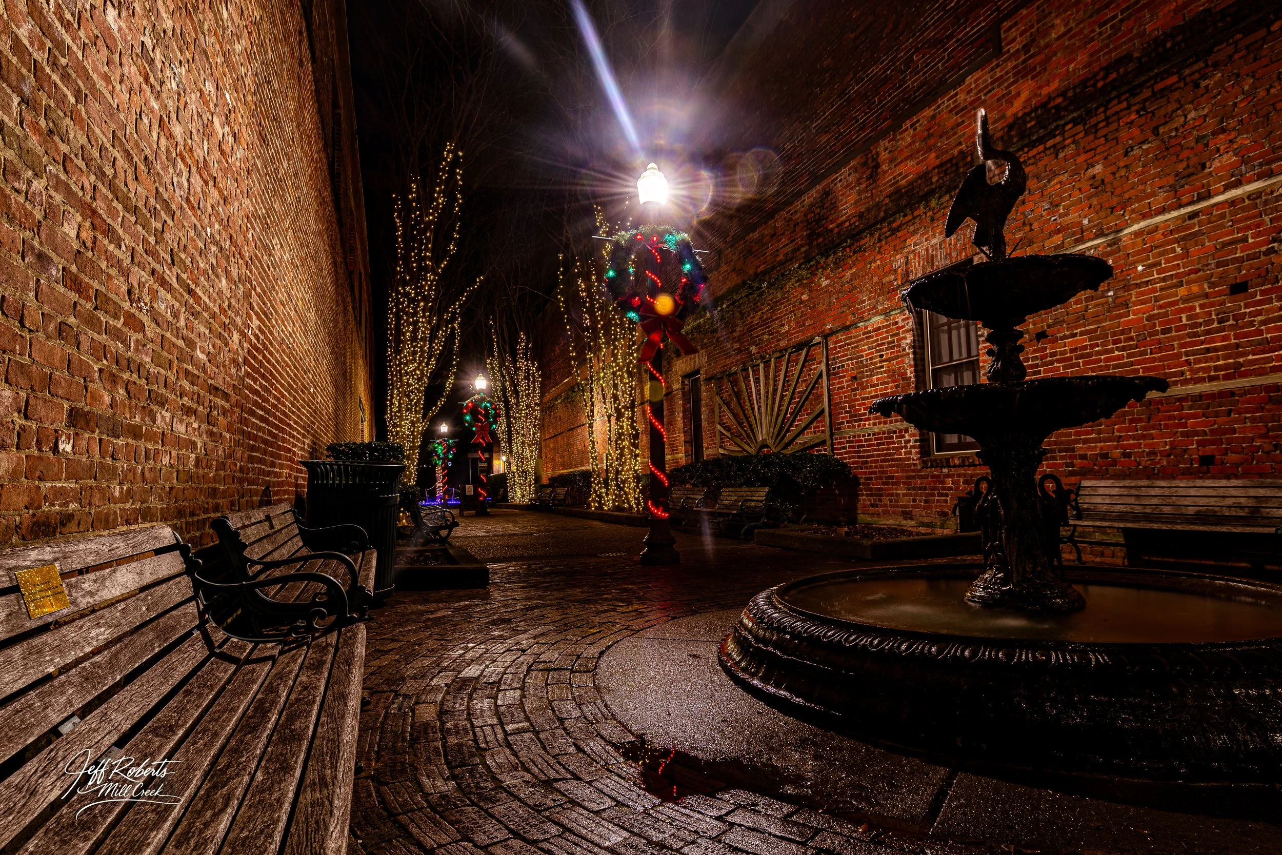 Nighttime scene of an outdoor brick walkway decorated for Christmas, with benches, a fountain, lit trees, and Christmas wreaths with lights and ribbons.