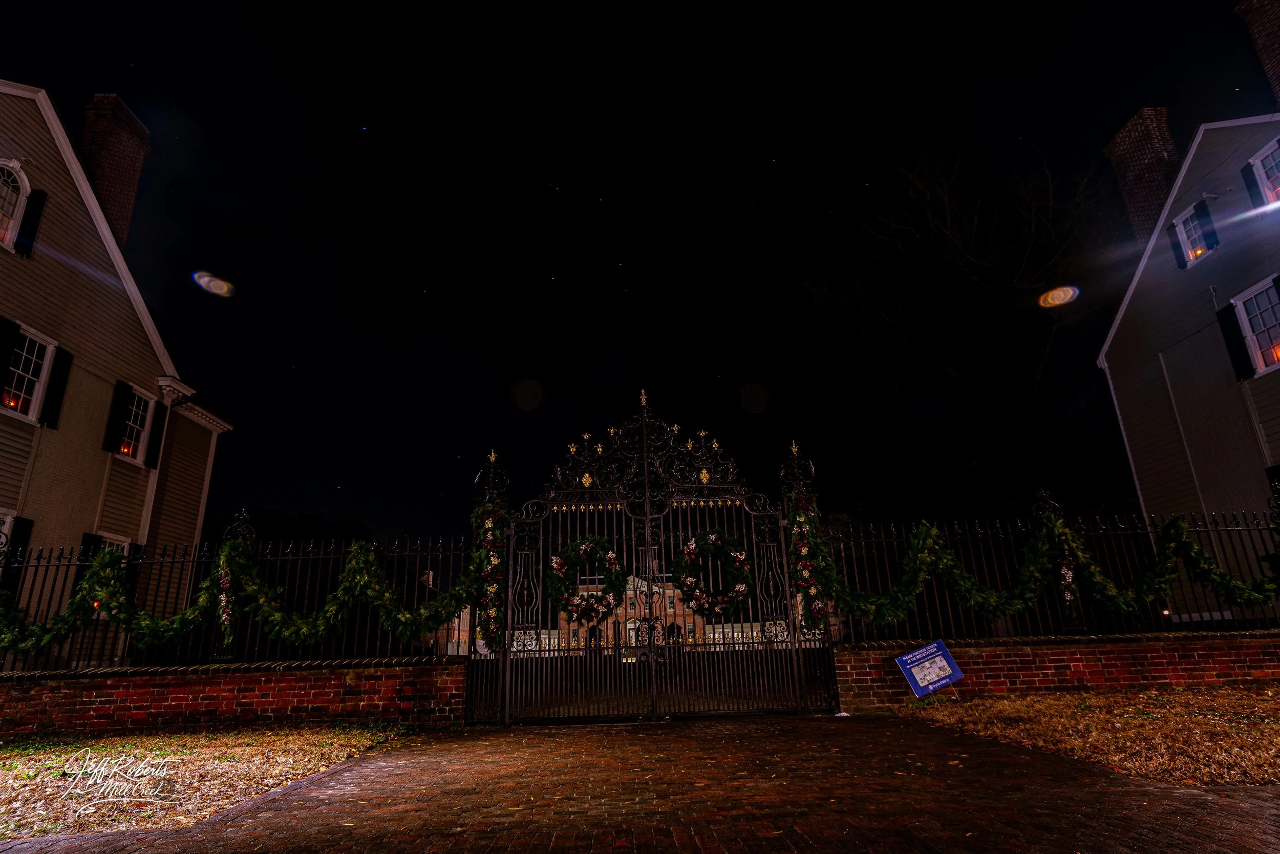 Nighttime view of an ornate metal gate decorated with Christmas garlands and wreaths, flanked by two large buildings, under a starry sky with lens flares.
