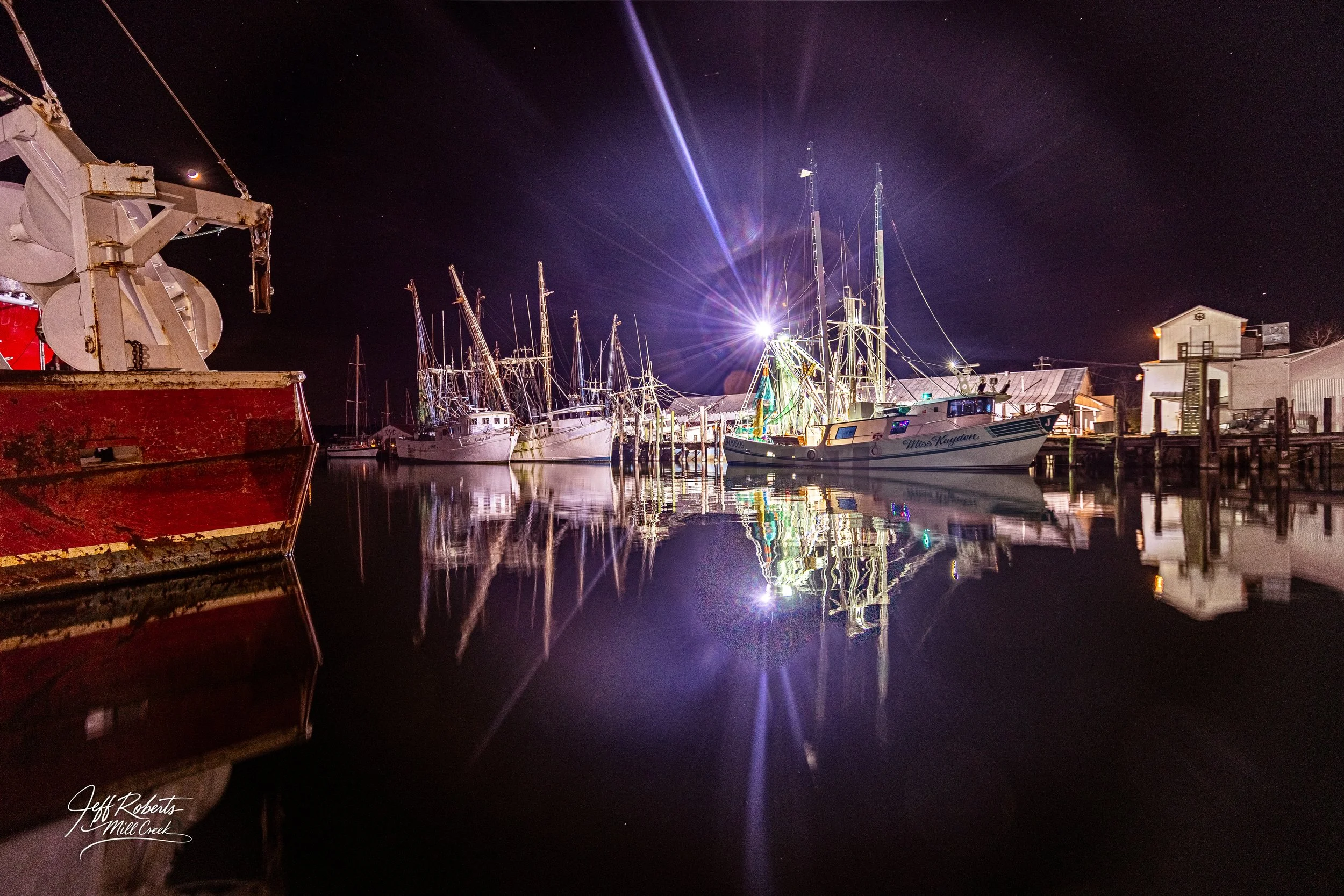 Nighttime view of sailboats and a fishing boat docked at a marina, with reflections on calm water and a bright light illuminating the scene.