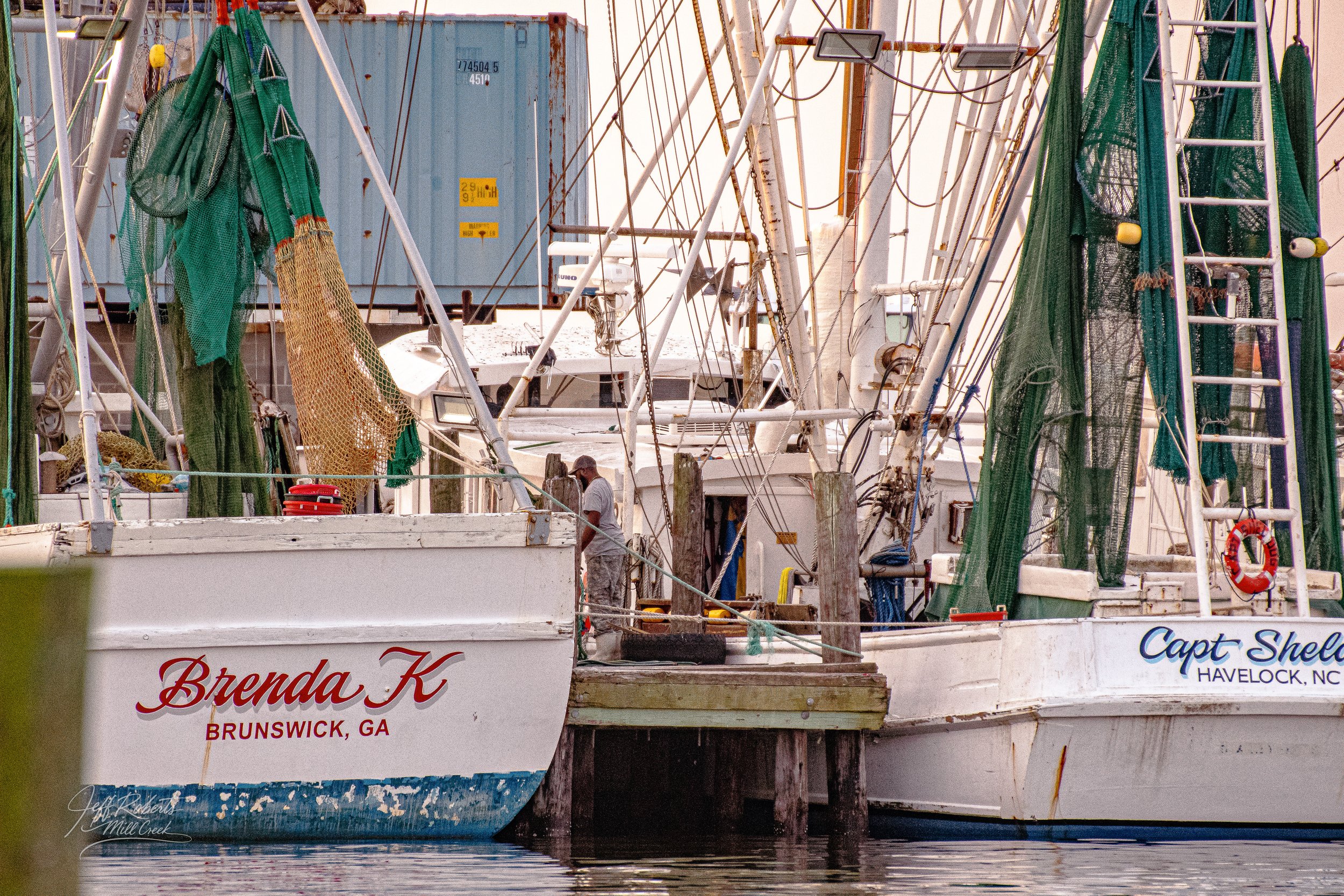 Two sailboats docked at a marina with fishing gear on the boats, including nets and rods. One boat has the name 'Brenda K' with Brunswick, GA written below. The other boat has the name 'Capt Shel' with Havelock, NC written below. There are wooden pos