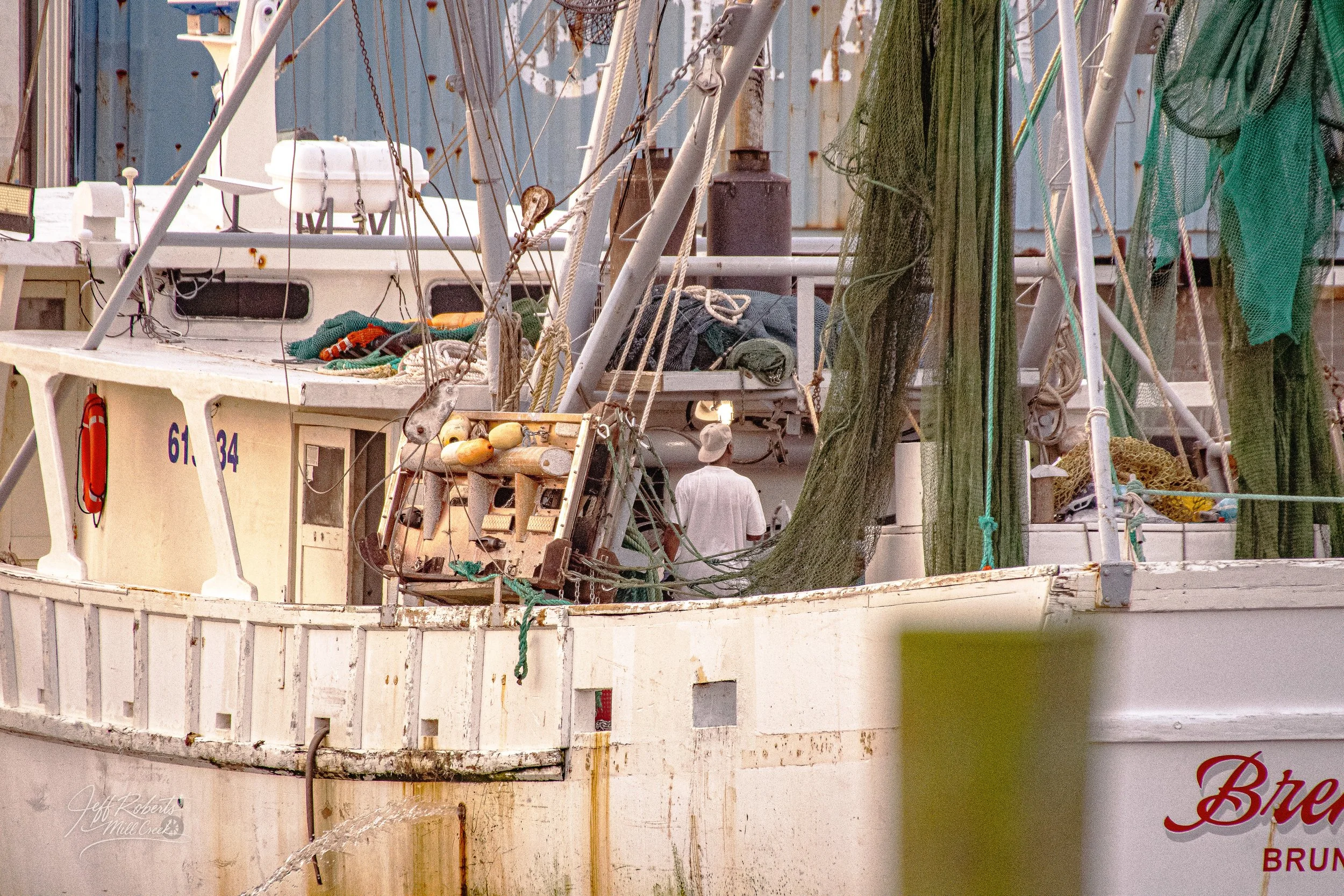 A boat docked at a marina with fishing gear and nets hanging on its sides. A person in a white shirt and hat is seen in the background.