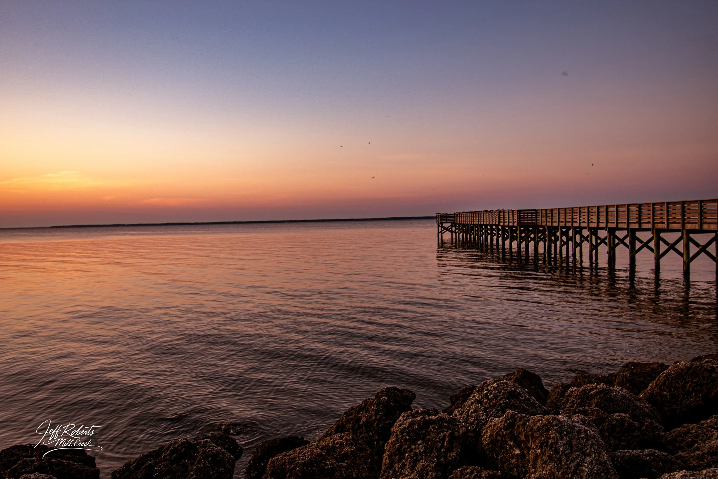 Sunset over a calm body of water with rocks in the foreground and a wooden pier extending into the water on the right. The sky is a gradient of soft pink, purple, and orange hues.