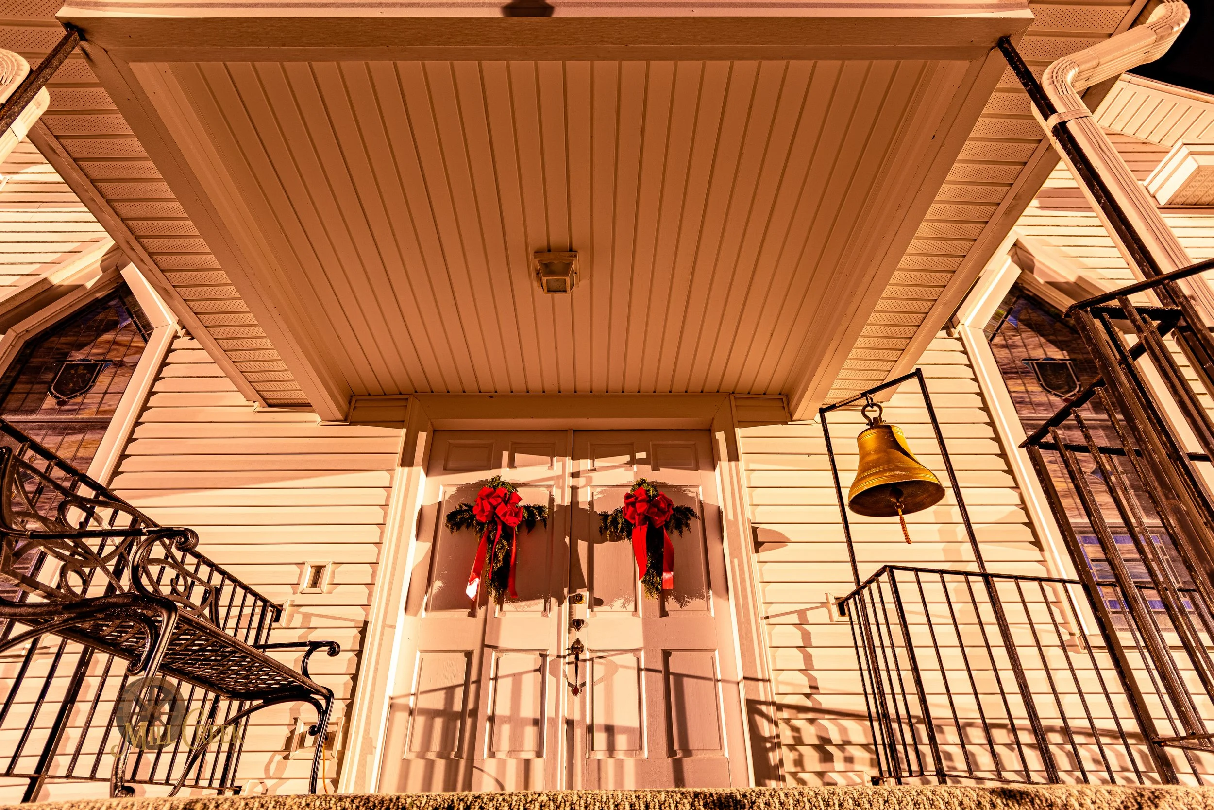 Front porch of a house decorated with holiday wreaths and red bows on the door, with a brass bell hanging on the side, black wrought iron railing, and yellow siding under a large roof.