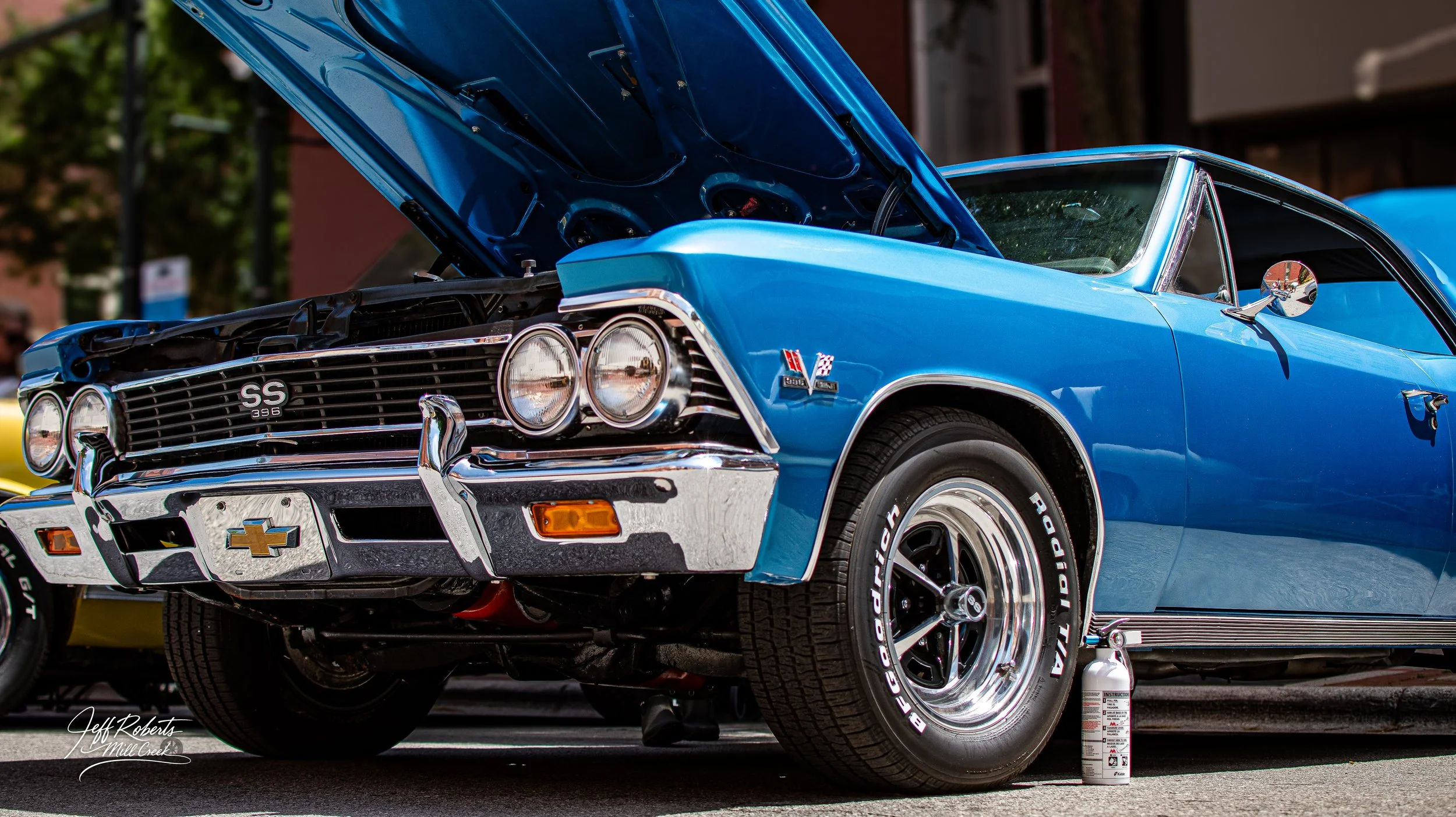 Blue vintage Chevrolet muscle car with open hood at car show, chrome details, rally tires, and fire extinguisher on the ground nearby.