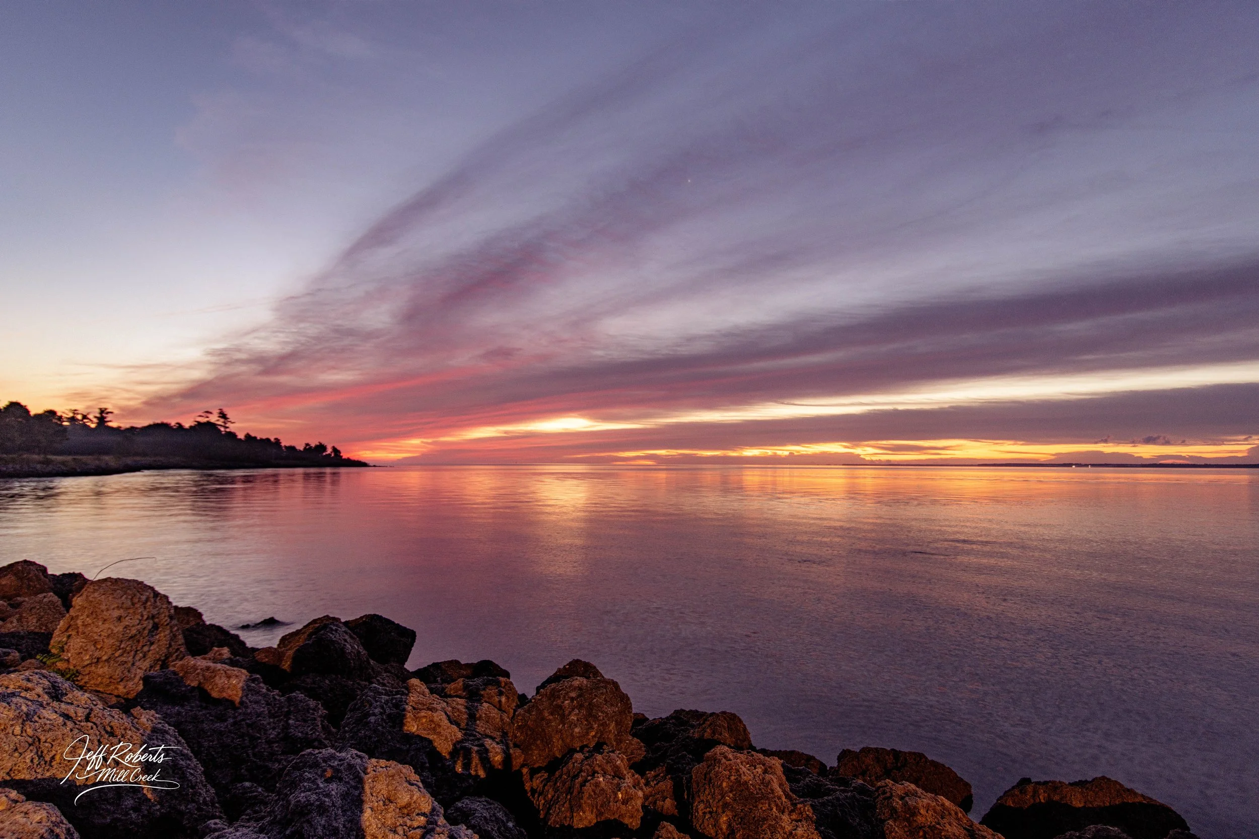 Sunset over a calm body of water with rocky shoreline in the foreground and a colorful sky with clouds in shades of purple, pink, orange, and yellow.