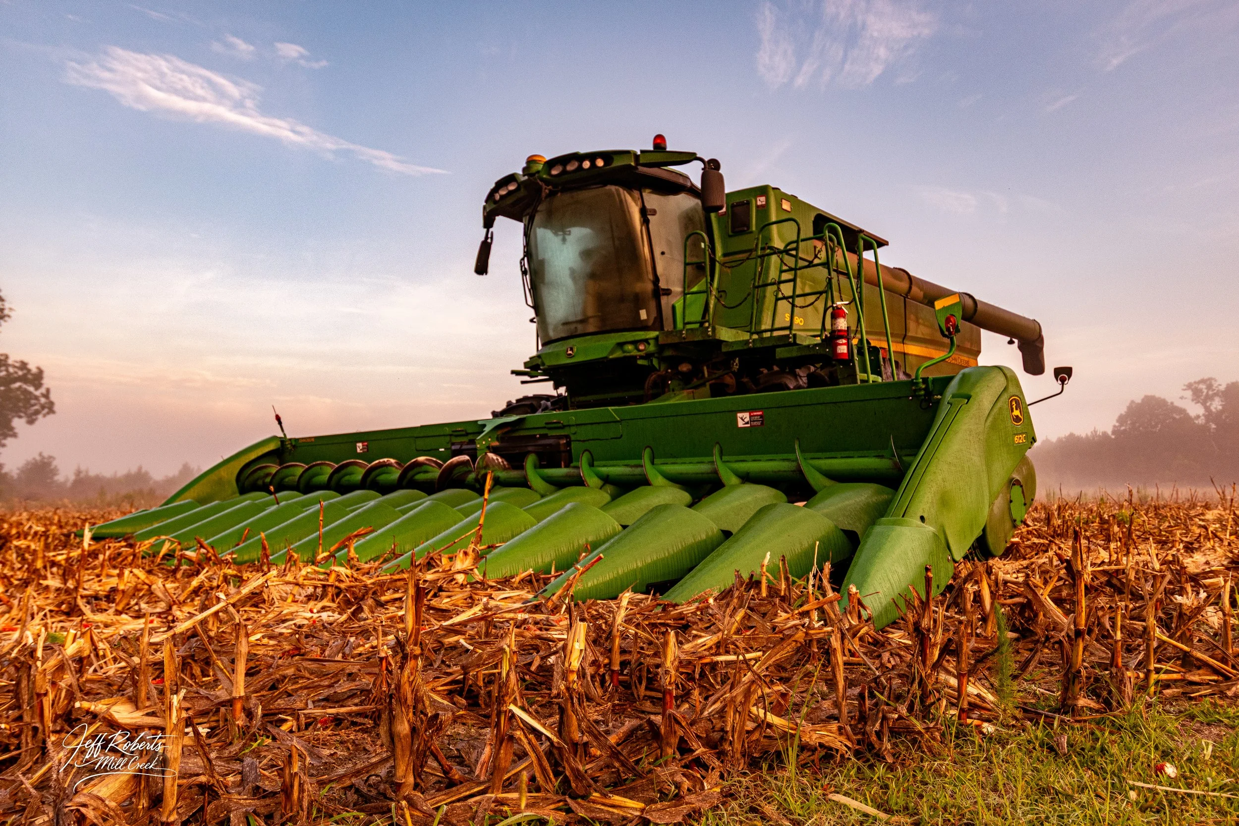A large green John Deere combine harvester working in a cornfield during sunset, with the sky filled with scattered clouds and trees on the horizon.