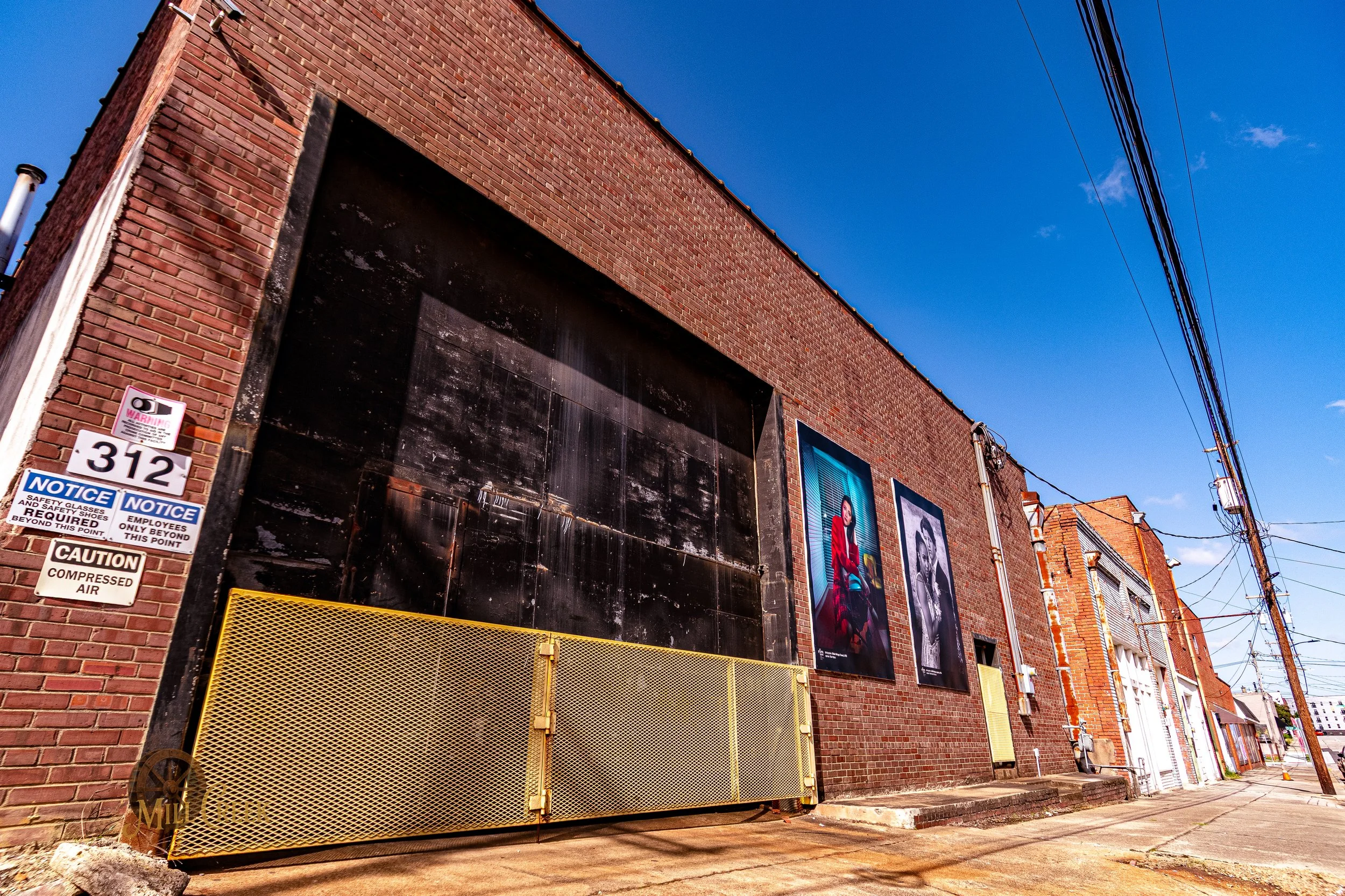 A brick building with two large posters of a woman in red and a man in a jacket on the outside wall. The building has a large black section and a yellow gate in front. There are street signs on the left and power lines overhead. The sky is clear and 