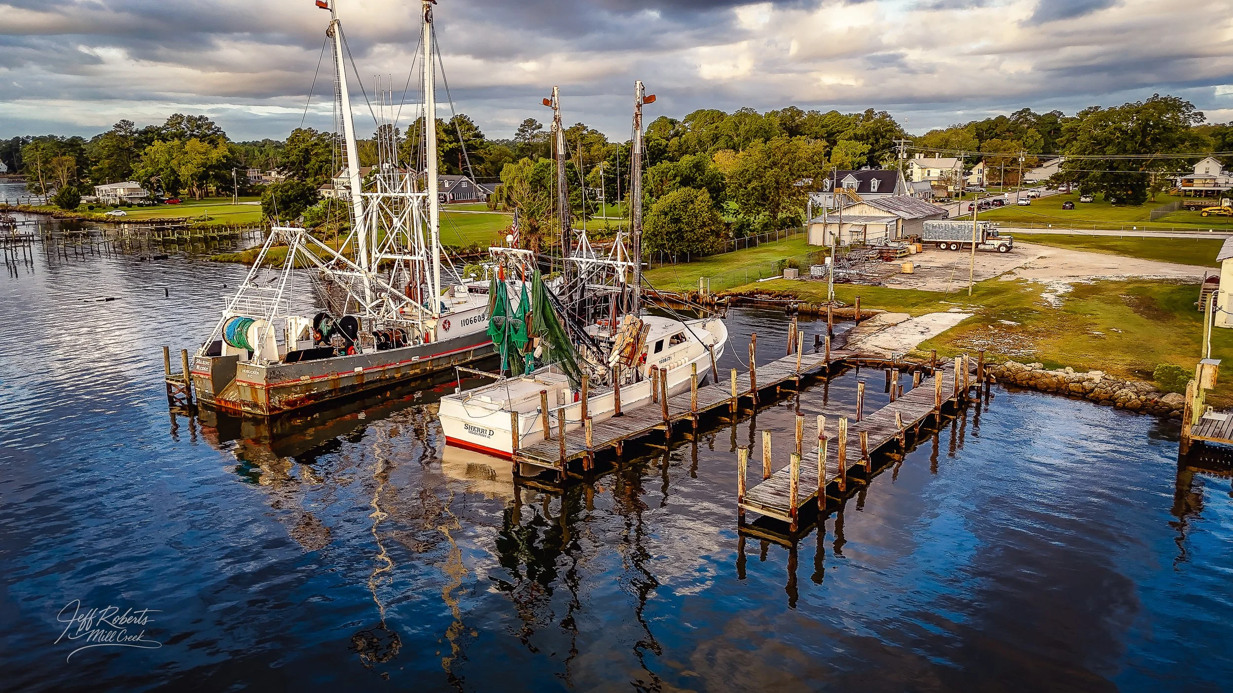 Aerial view of two boats docked at a marina, with residential houses, trees, and cloudy sky in the background.