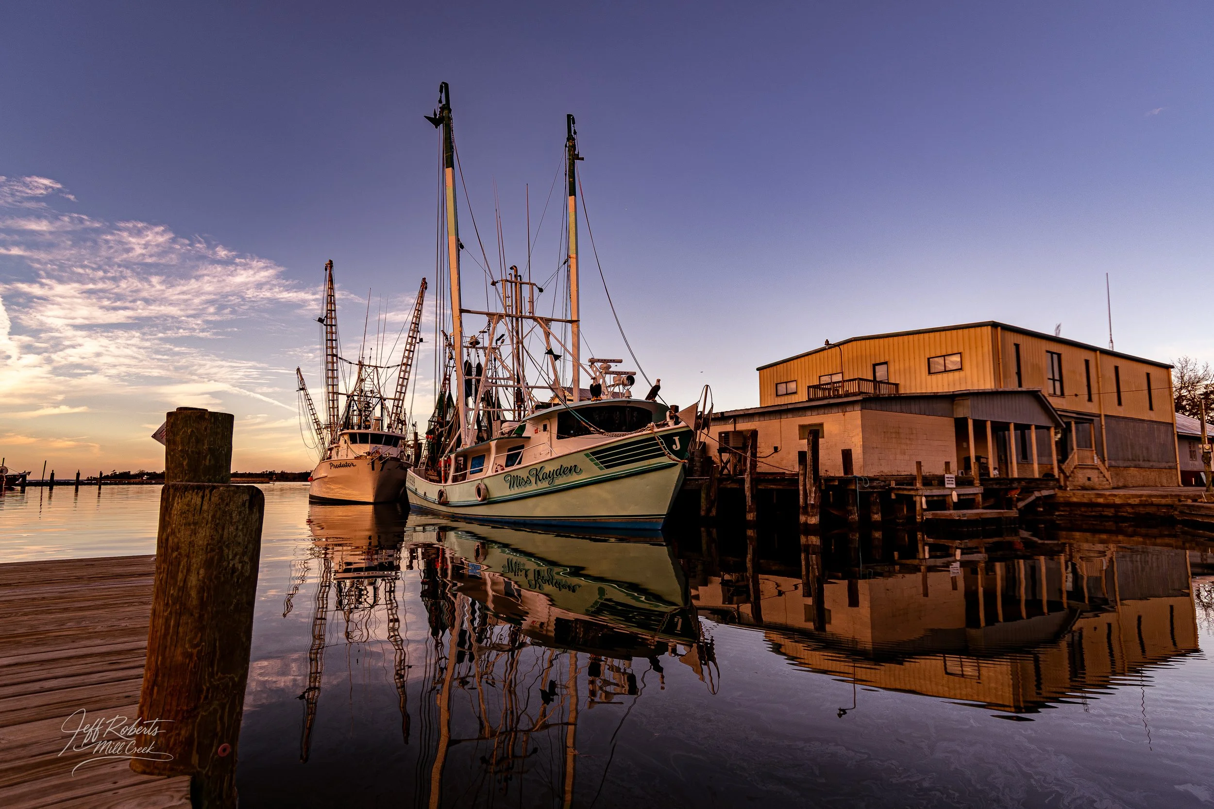Boats docked along a pier at sunset with calm water and a partly cloudy sky.