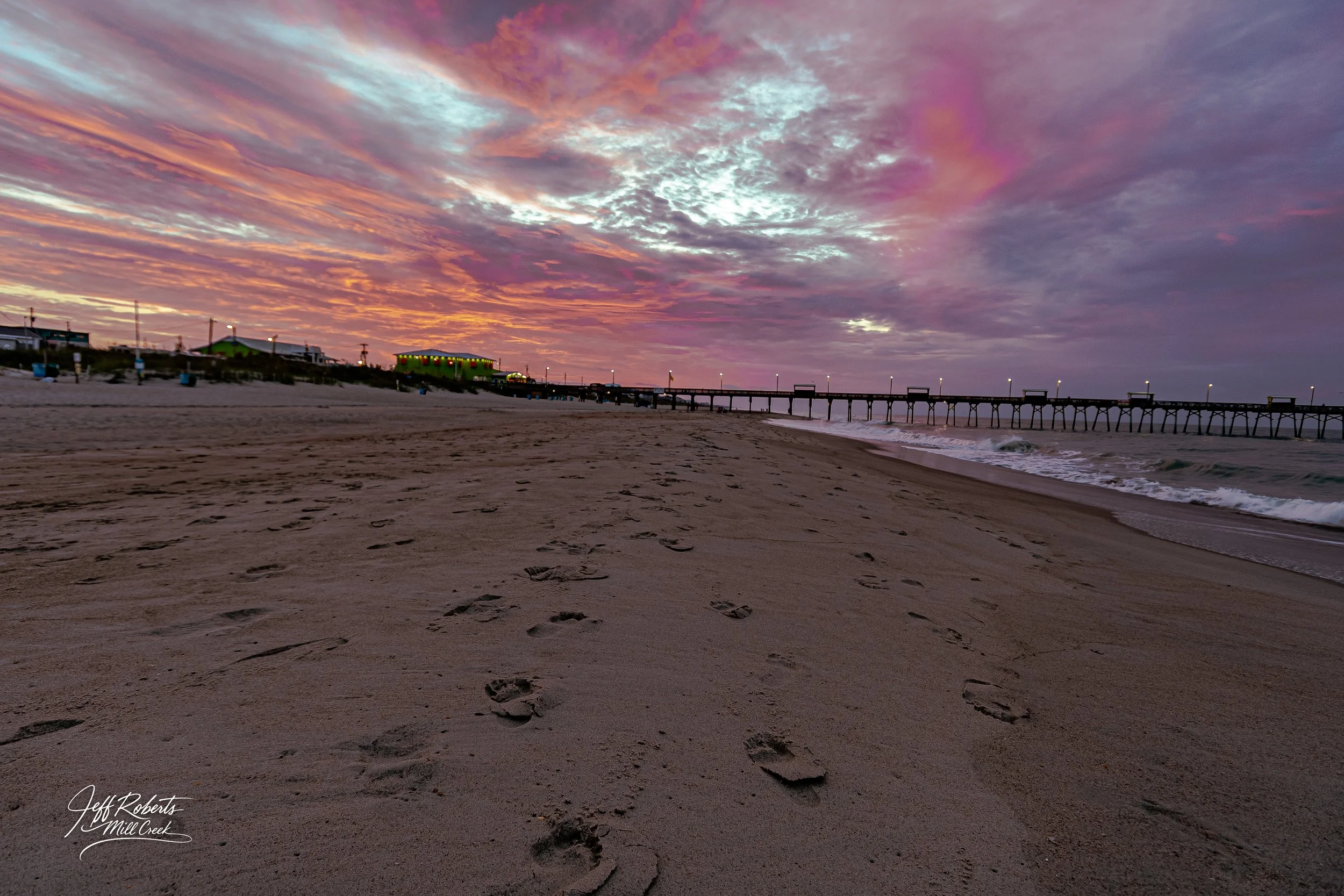 Footprints in the sand on a beach at sunset with a pier extending into the water and colorful clouds in the sky.