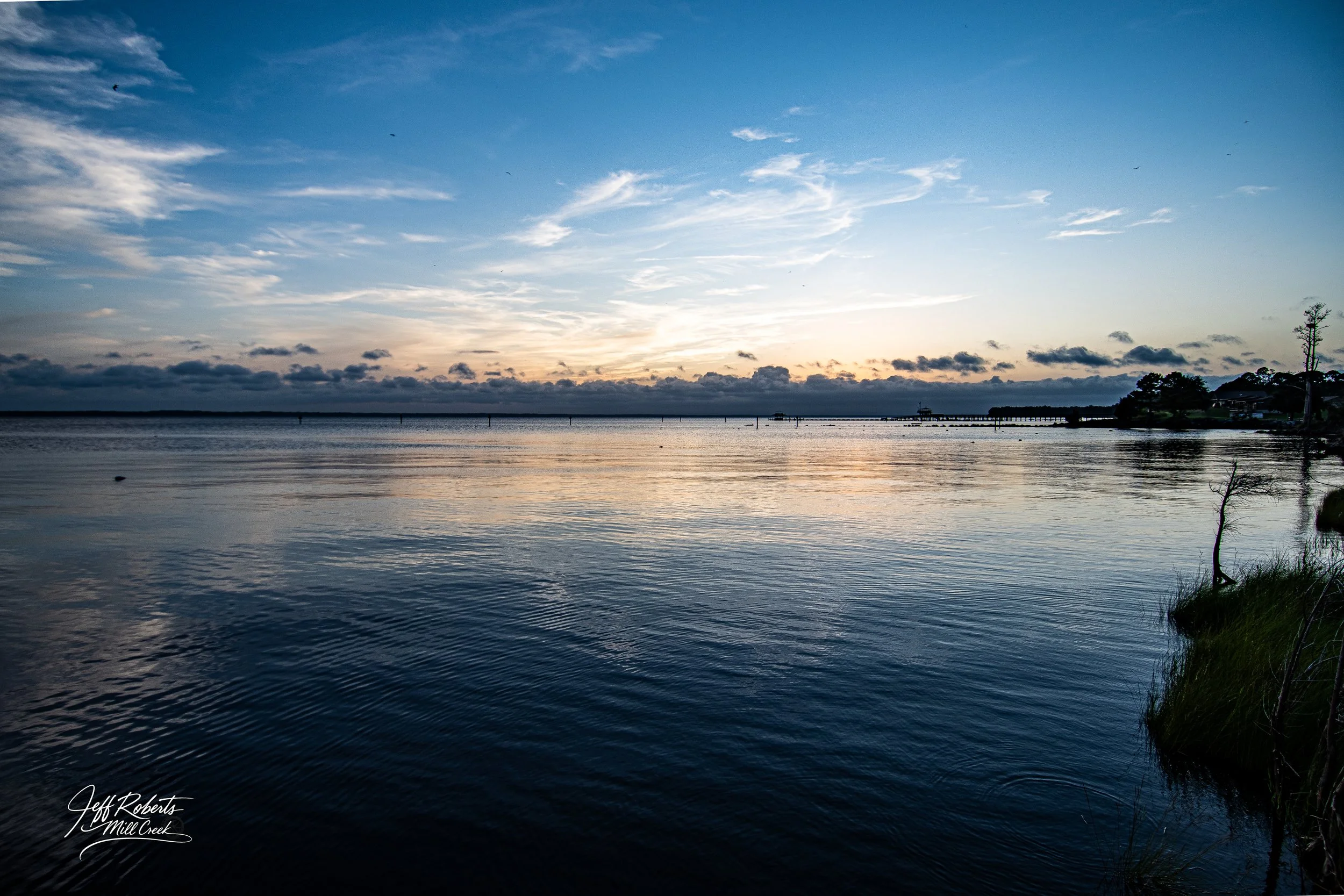 Sunset over a calm body of water with a partly cloudy sky and distant trees and structures on the horizon.