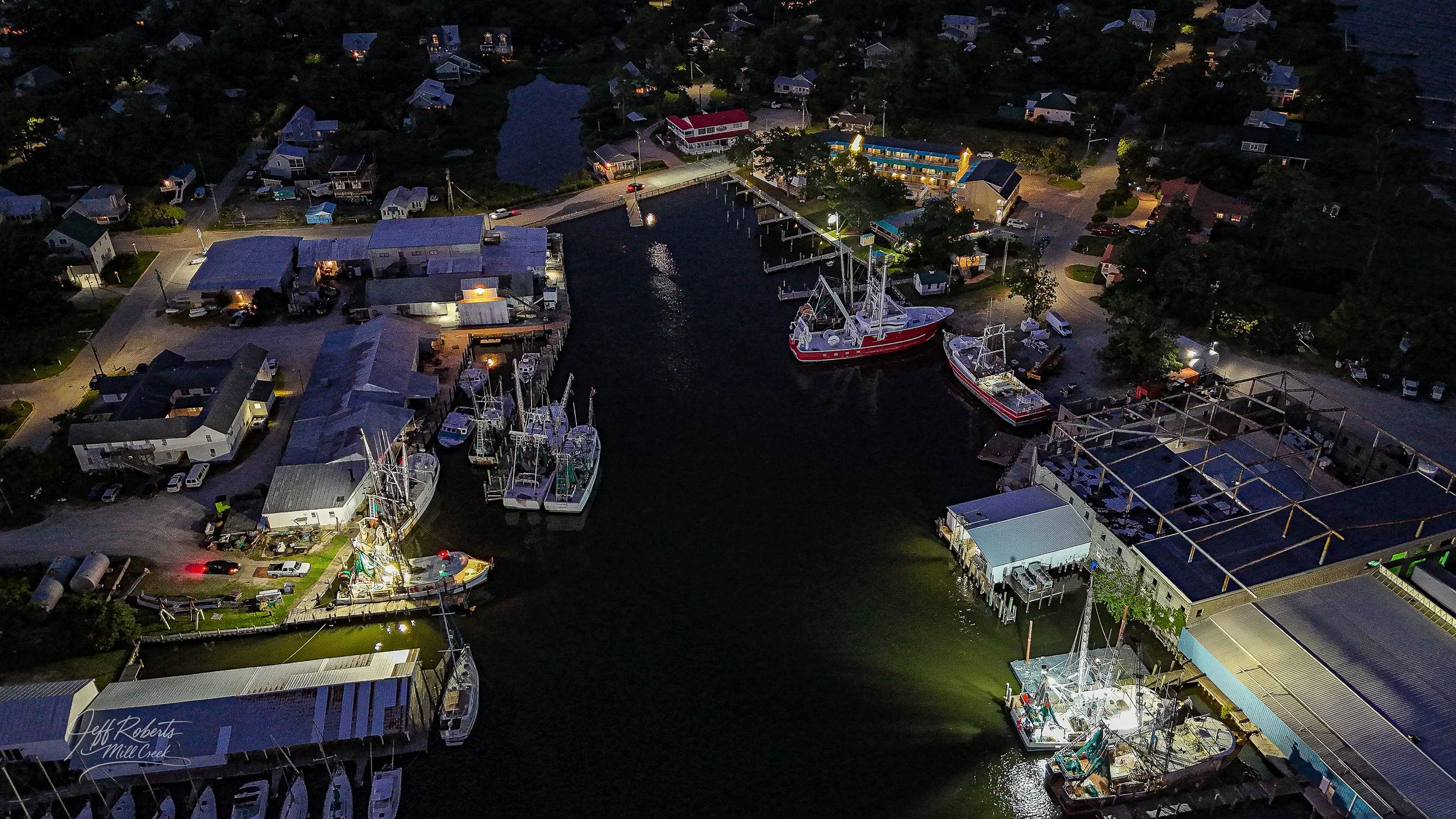 Bird's eye view of a marina at night with boats docked in the water and illuminated buildings along the shore.
