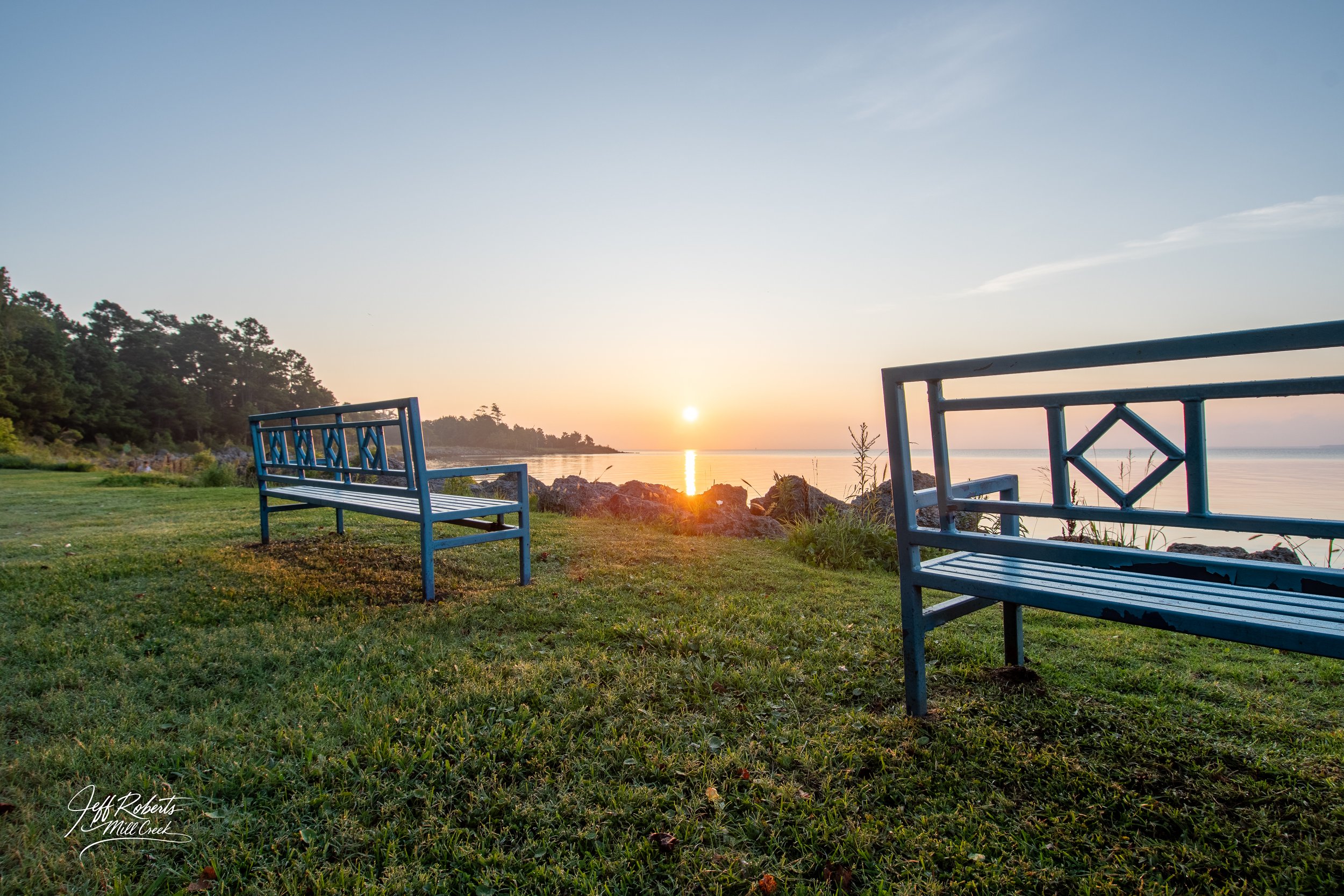 Sunset over a calm body of water with two blue benches on a grassy area in the foreground, trees on the left side, rocks near the shore, and a clear sky.
