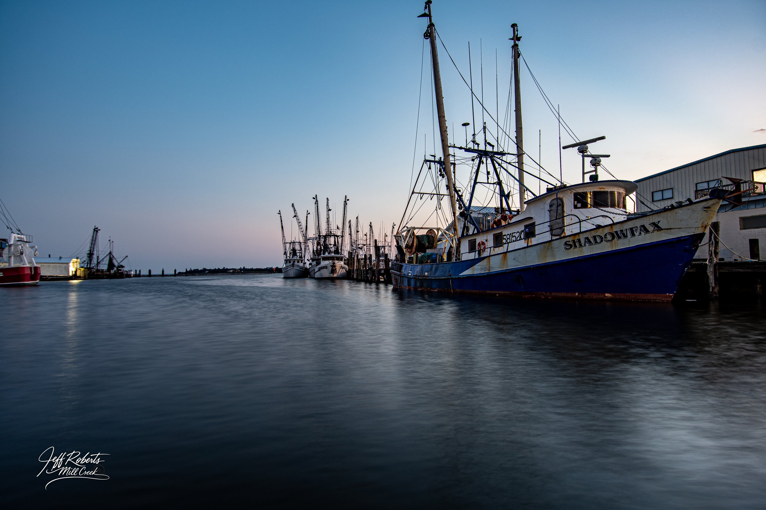 Several fishing boats docked at a harbor during dusk, with calm water and a clear sky.
