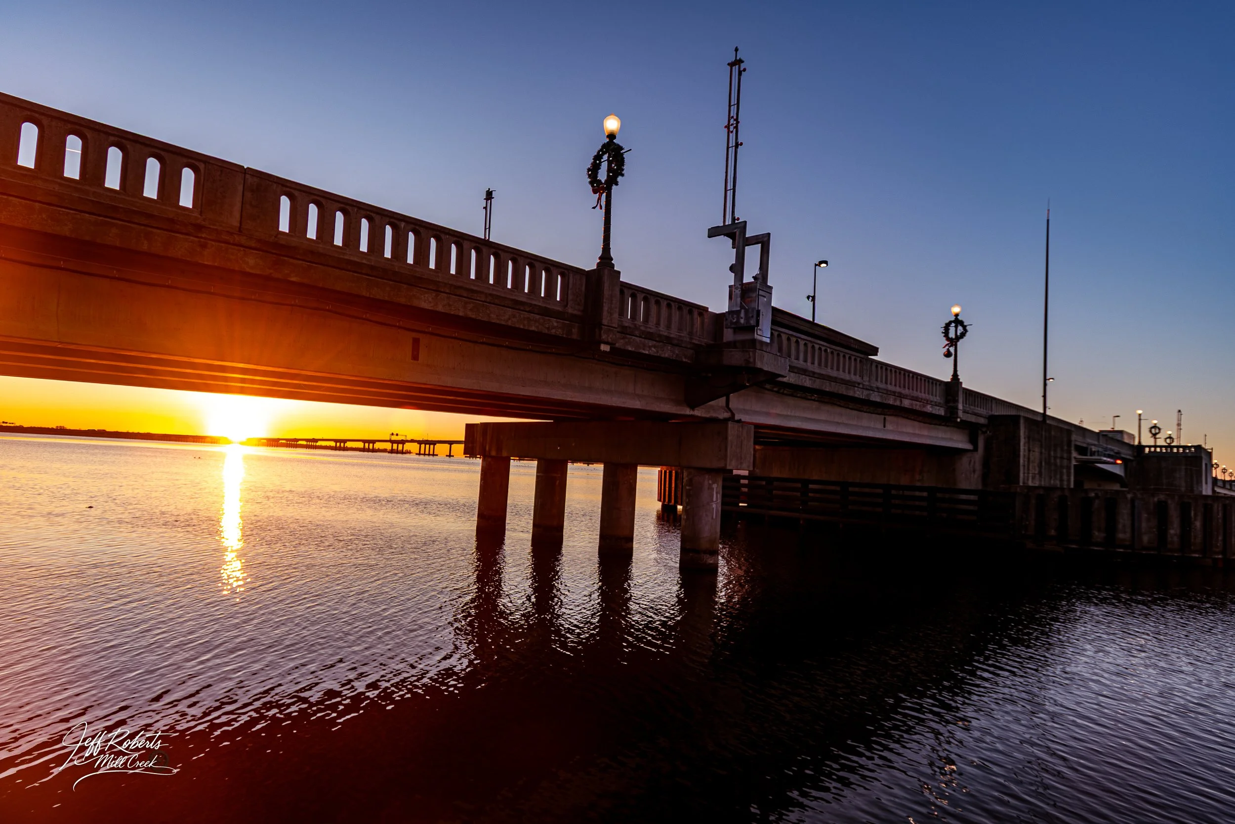 A bridge over a body of water at sunset, with the sun near the horizon and reflections on the water, decorated with holiday wreaths on the lamps.