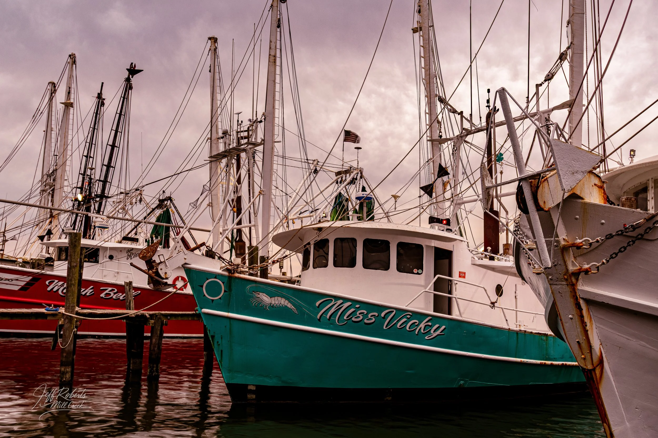 A marina with multiple docked boats, including a teal boat named Miss Vicky, with masts and rigging against a cloudy sky.