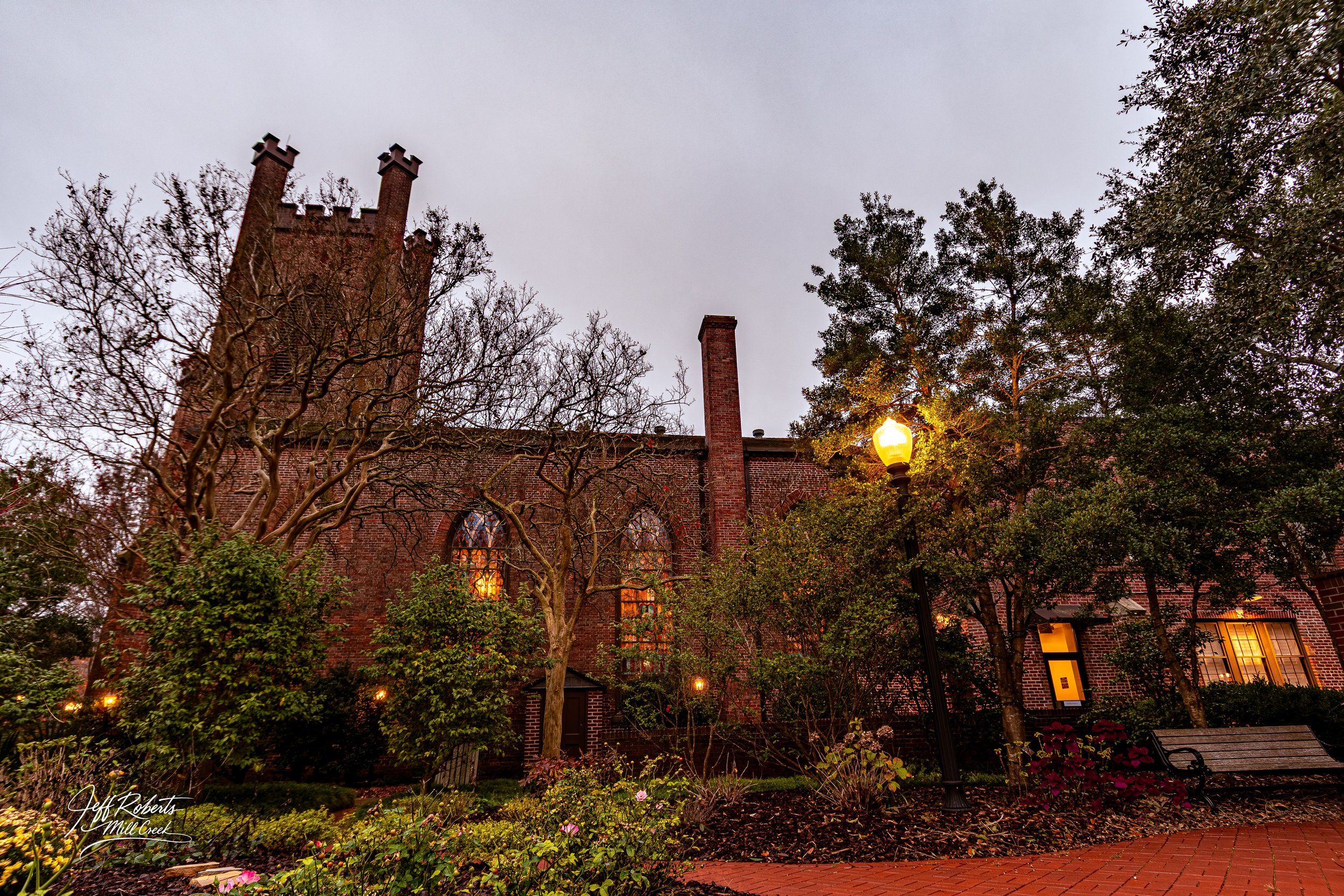 An old brick church or building with Gothic windows, surrounded by trees with some leaves, a lit street lamp, a brick pathway, and a wooden bench in the foreground during dusk.