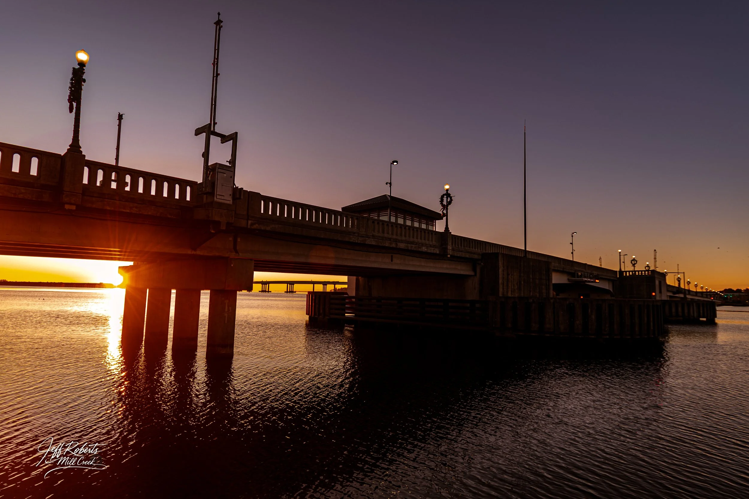 A pier extending into a body of water at sunset, with lamp posts along the railings and a small building on the pier. The sun is setting on the horizon, casting an orange glow across the water and sky.