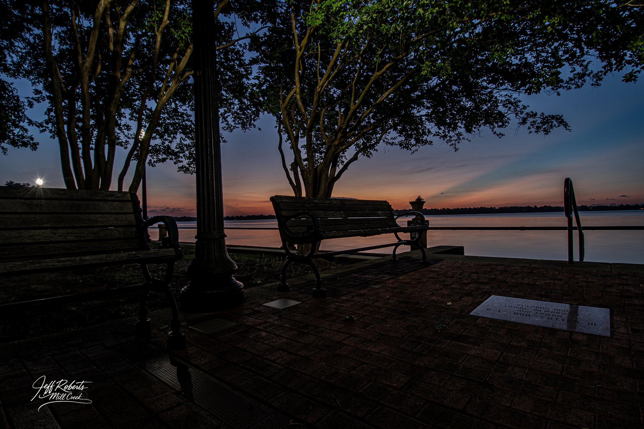 A lakeside park at dusk with two empty wooden benches, a large tree, a lamppost, and a staircase leading into the water, with the sky showing hues of orange and blue.