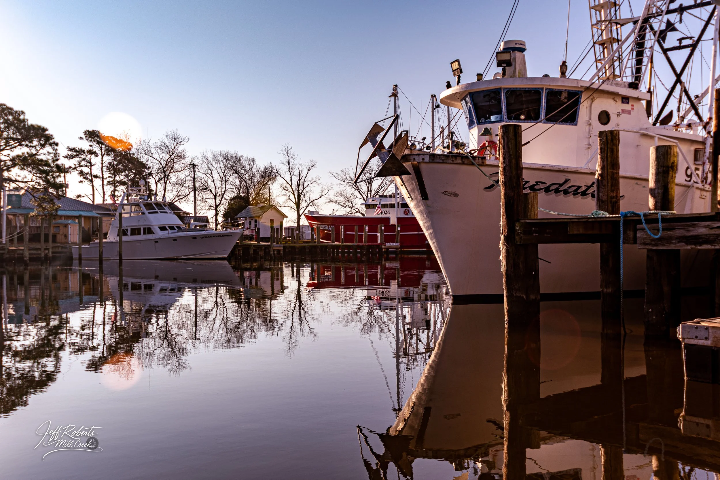 Boats docked at a marina during sunset with reflections on calm water, trees with no leaves, and small houses in the background.