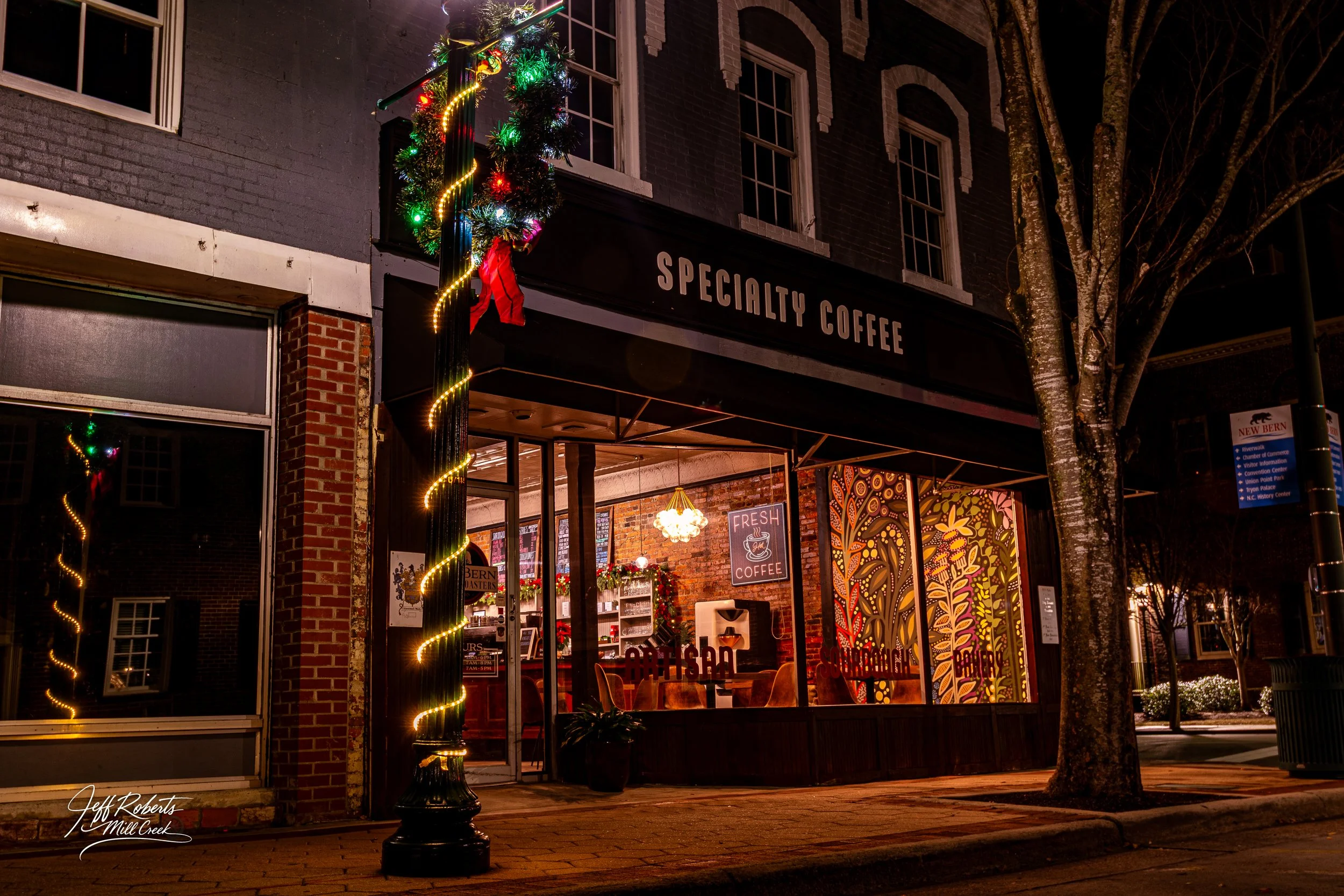 Nighttime view of a cozy coffee shop called 'Specialty Coffee' with decorated windows and a lit lamppost wrapped with Christmas lights and ornaments.