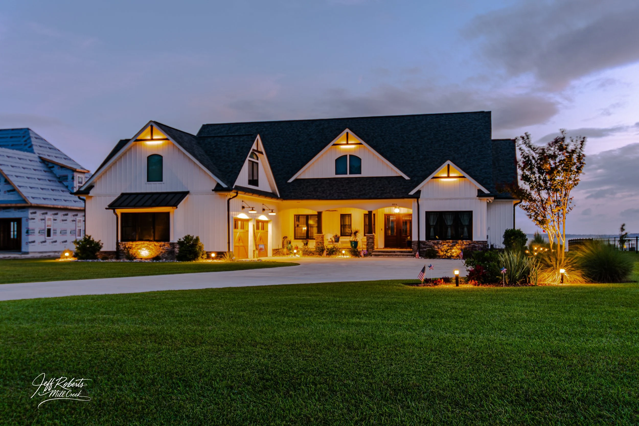 A modern two-story house with white exterior walls, a dark roof, and warm outdoor lighting at dusk. The house features multiple gables, a front porch with patio furniture, and a well-maintained lawn with landscaping. In the background, there is a nei