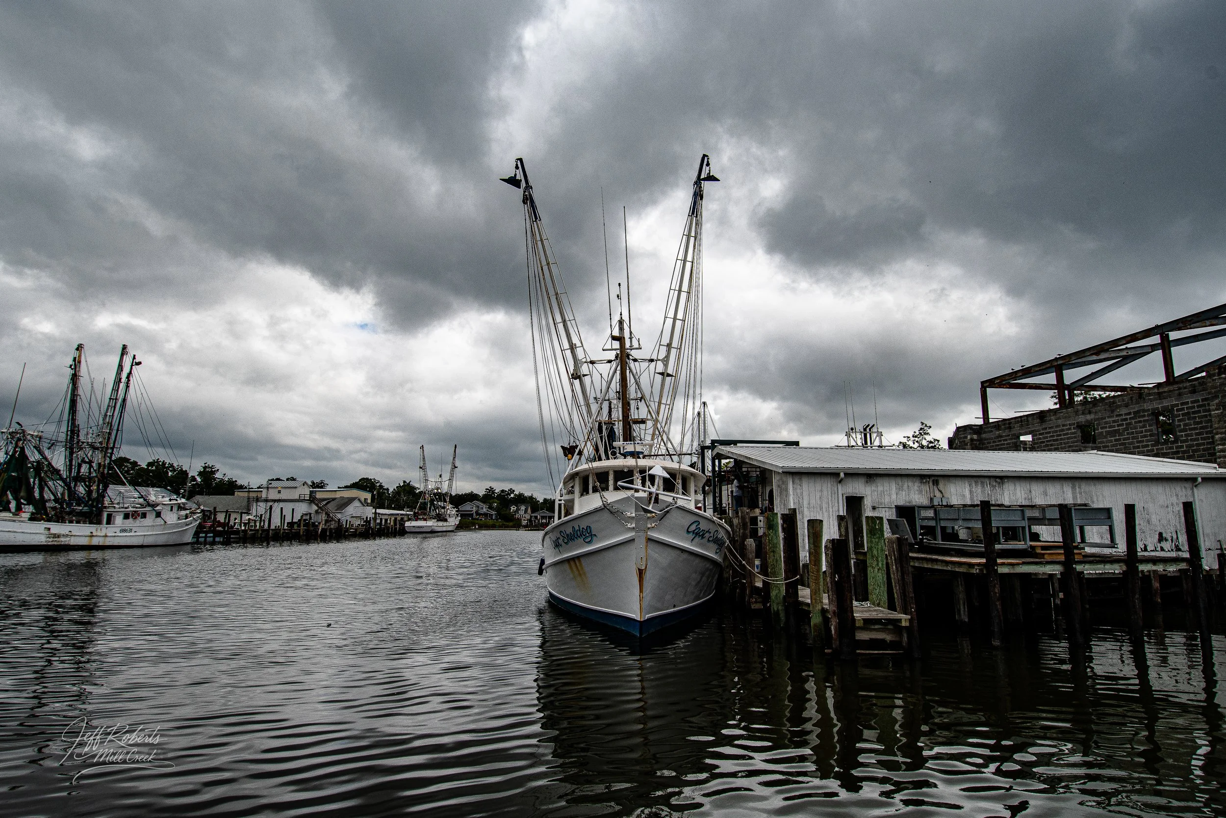 A boat docked at a pier on a cloudy day, with other boats visible in the background and dark, overcast sky.