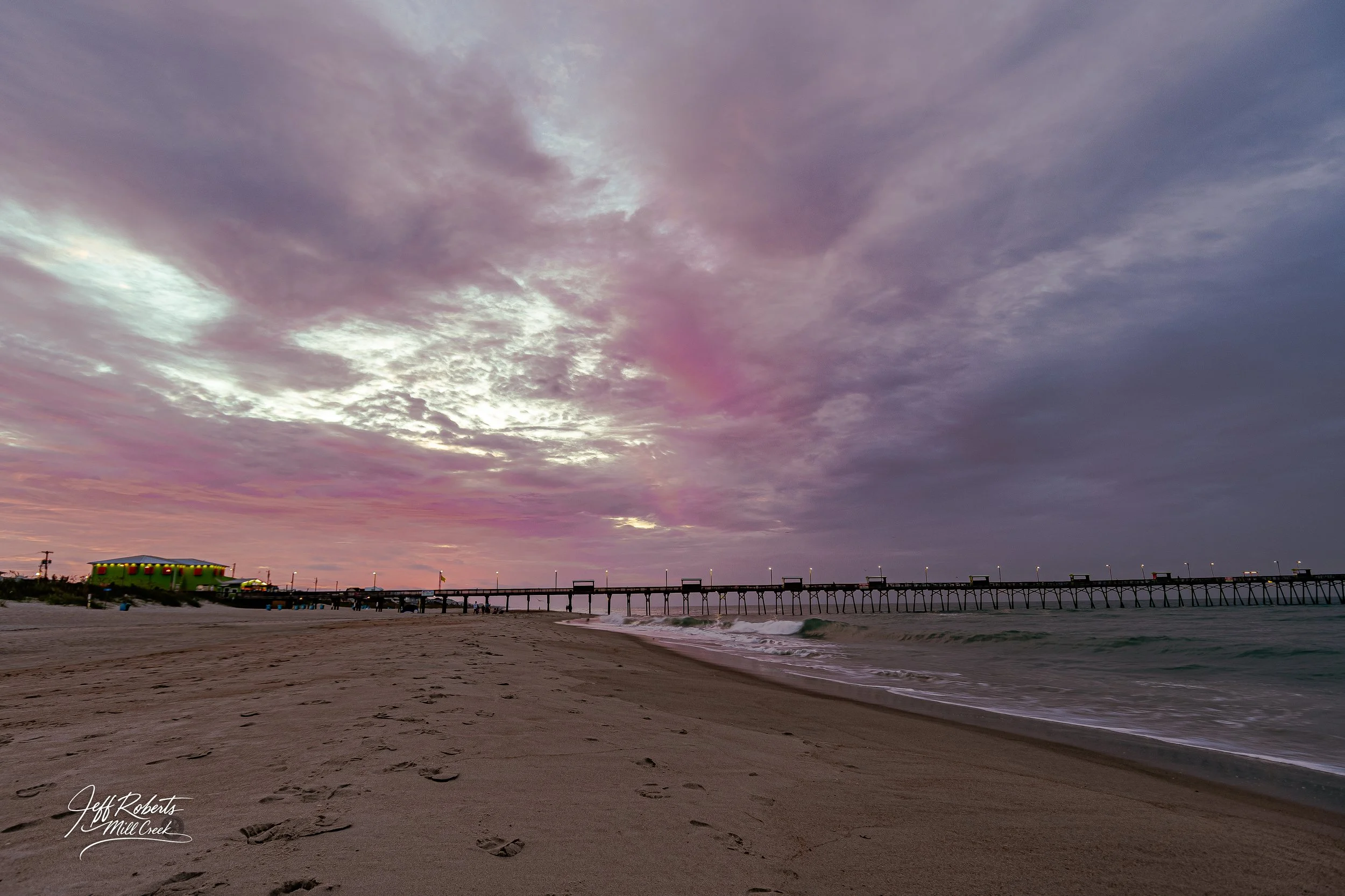 A sandy beach with footprints, ocean waves, a pier in the distance, and a cloudy sky with hints of pink and purple at sunset.