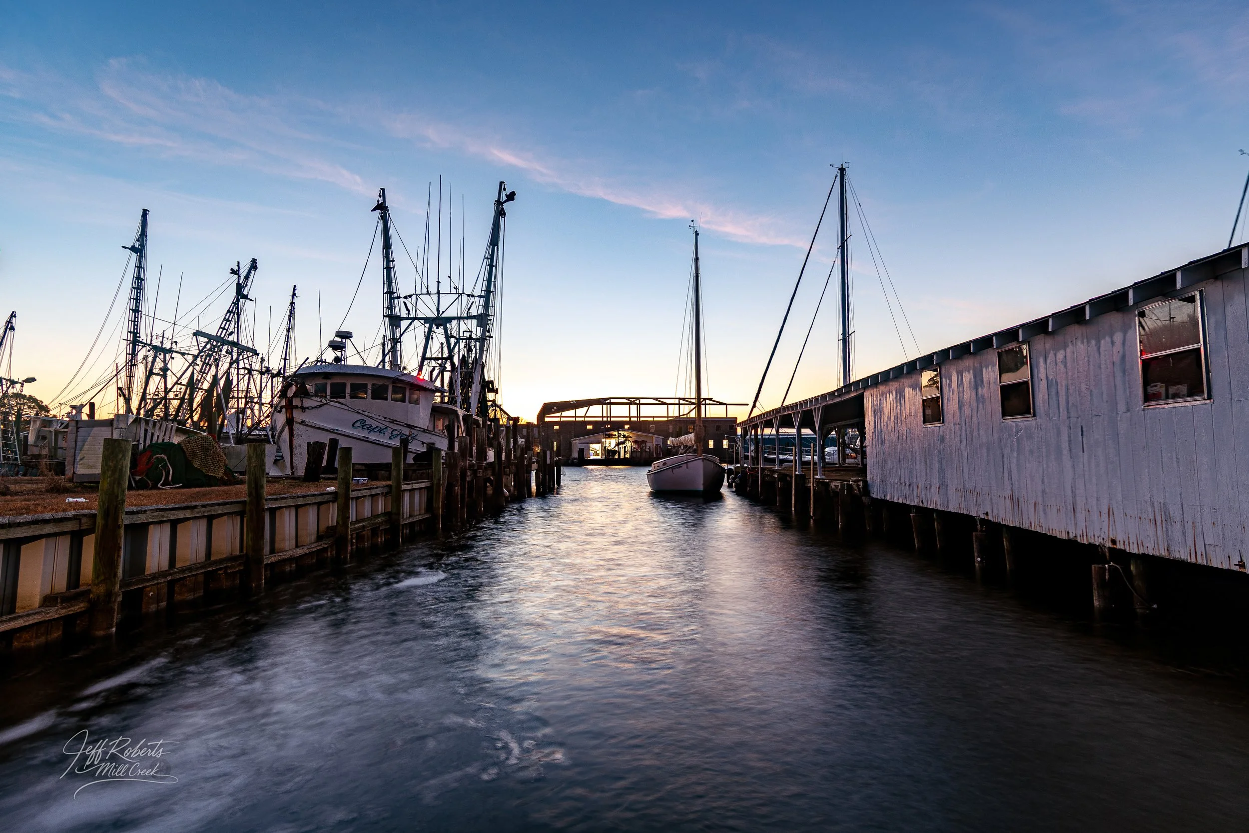 Boats docked at a marina during sunset with calm water, a clear sky, and a pier in the background.