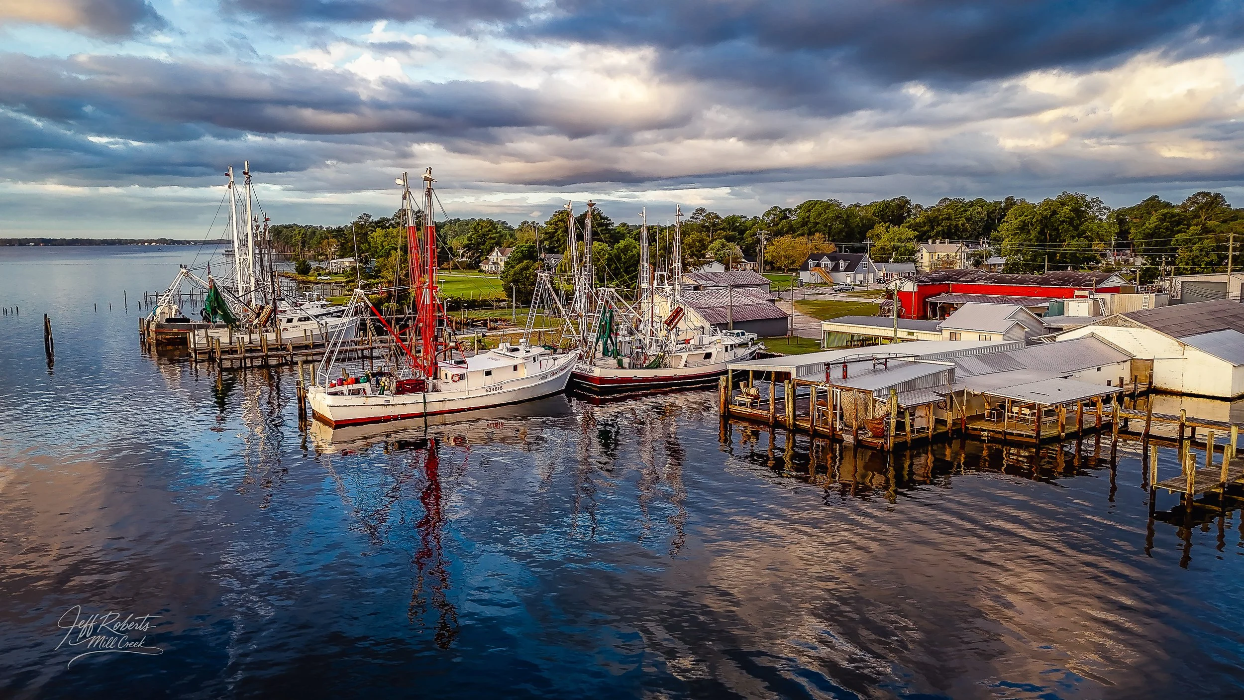 A small harbor with boats docked next to a wooden pier, surrounded by trees, houses, and buildings under a cloudy sky.