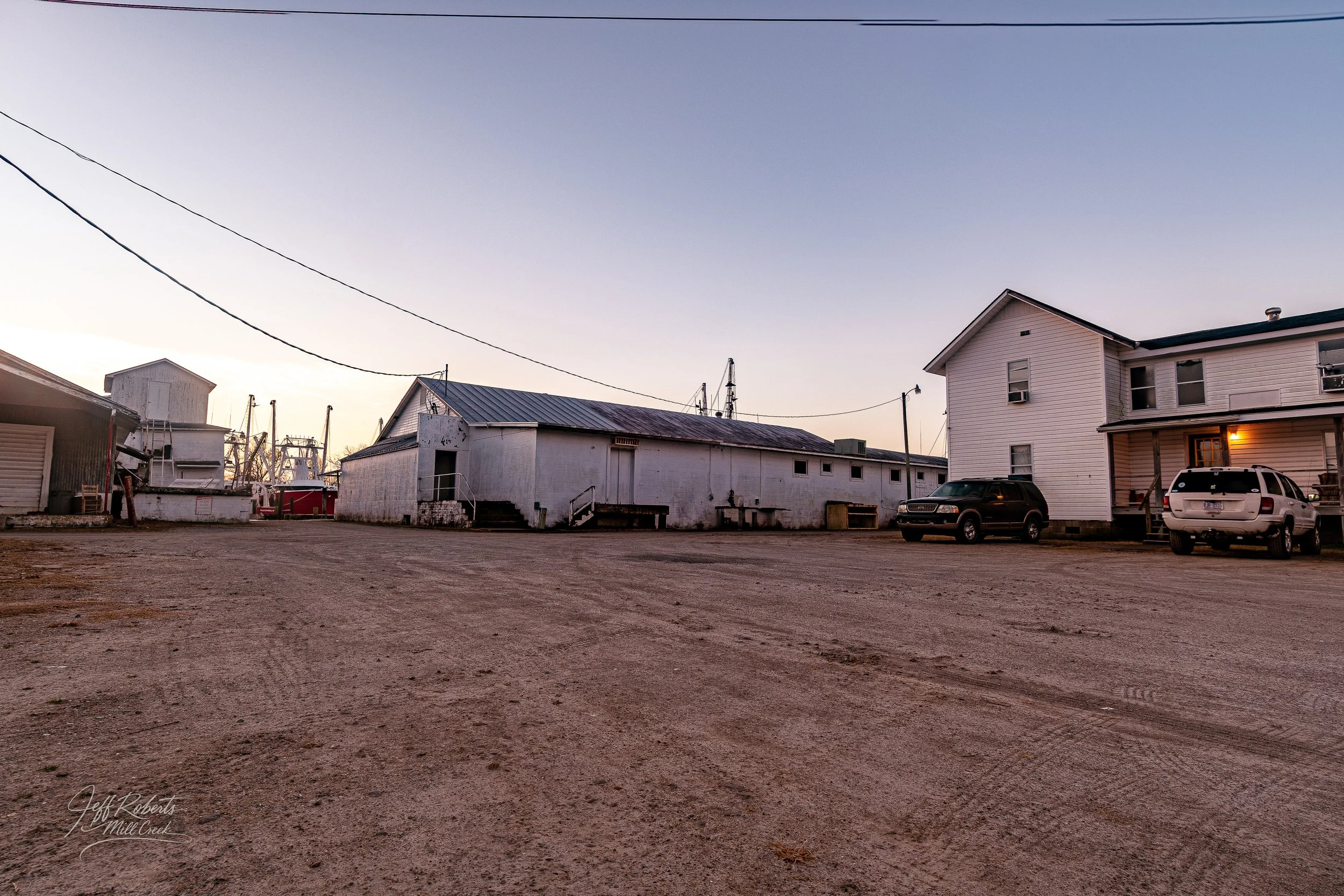 A dirt lot in front of white buildings and houses with cars parked in front, under a clear sky at sunset.