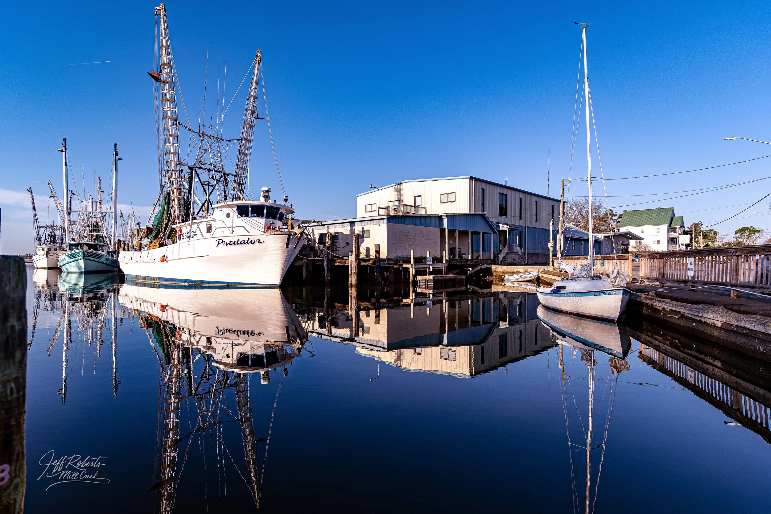Boats docked at a marina with calm water reflecting the boats and nearby buildings under a clear blue sky.